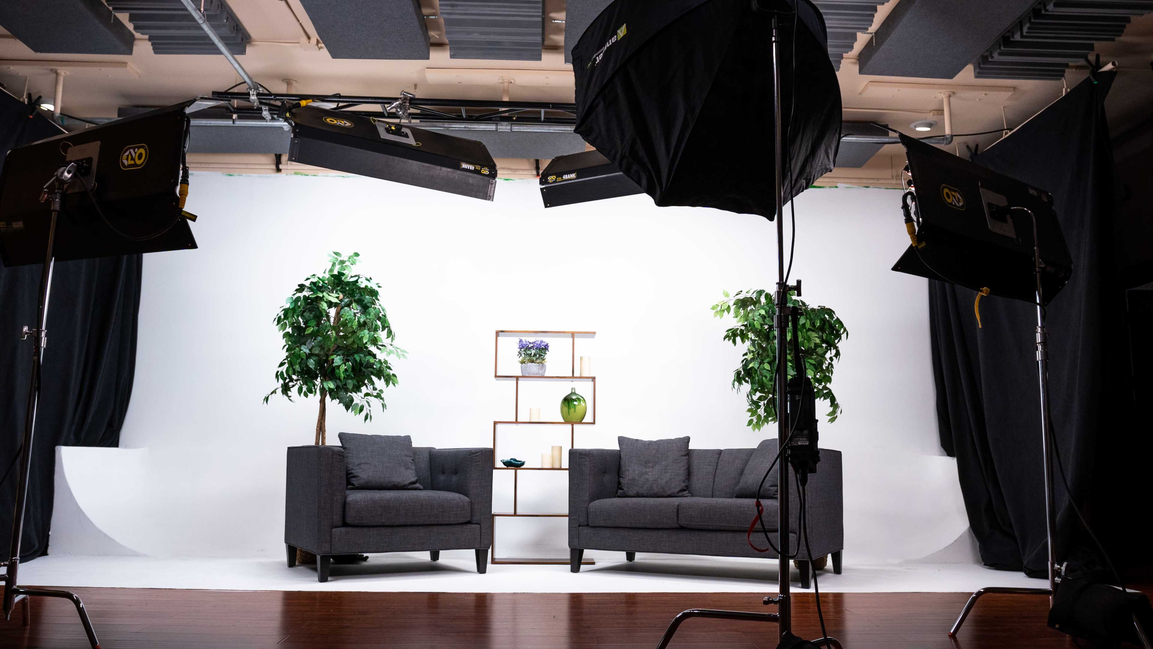 A photography studio setup featuring two gray sofas flanking a shelf with decorative items, surrounded by artificial plants and studio lighting.