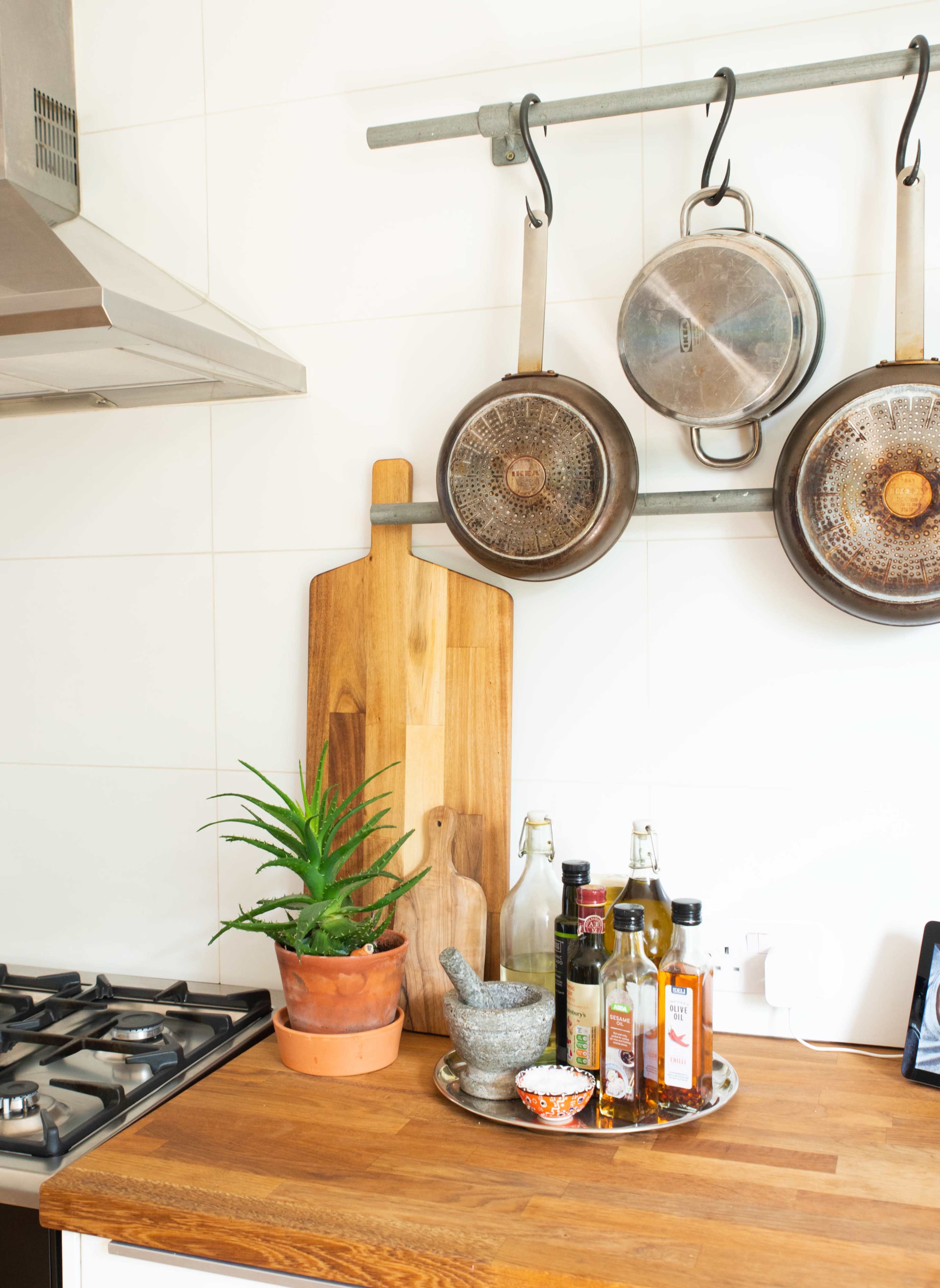 A kitchen counter features a wooden cutting board, a potted plant, various bottles of oils, and hanging pots on a metal rack.