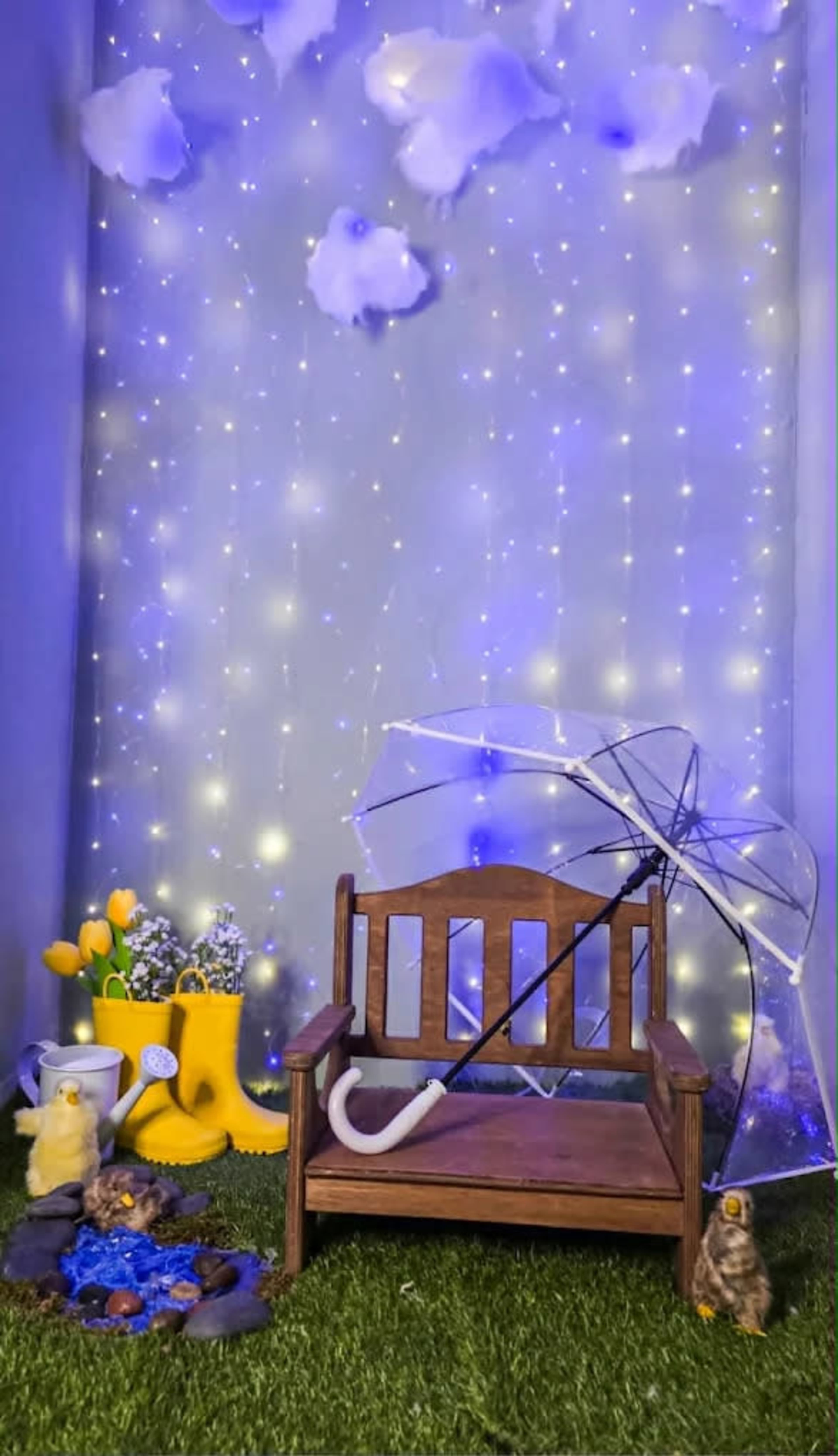 The image features a small wooden bench with a clear umbrella resting on it, surrounded by decorative elements like yellow rubber boots, blue flowers, and twinkling lights in a whimsical setting.