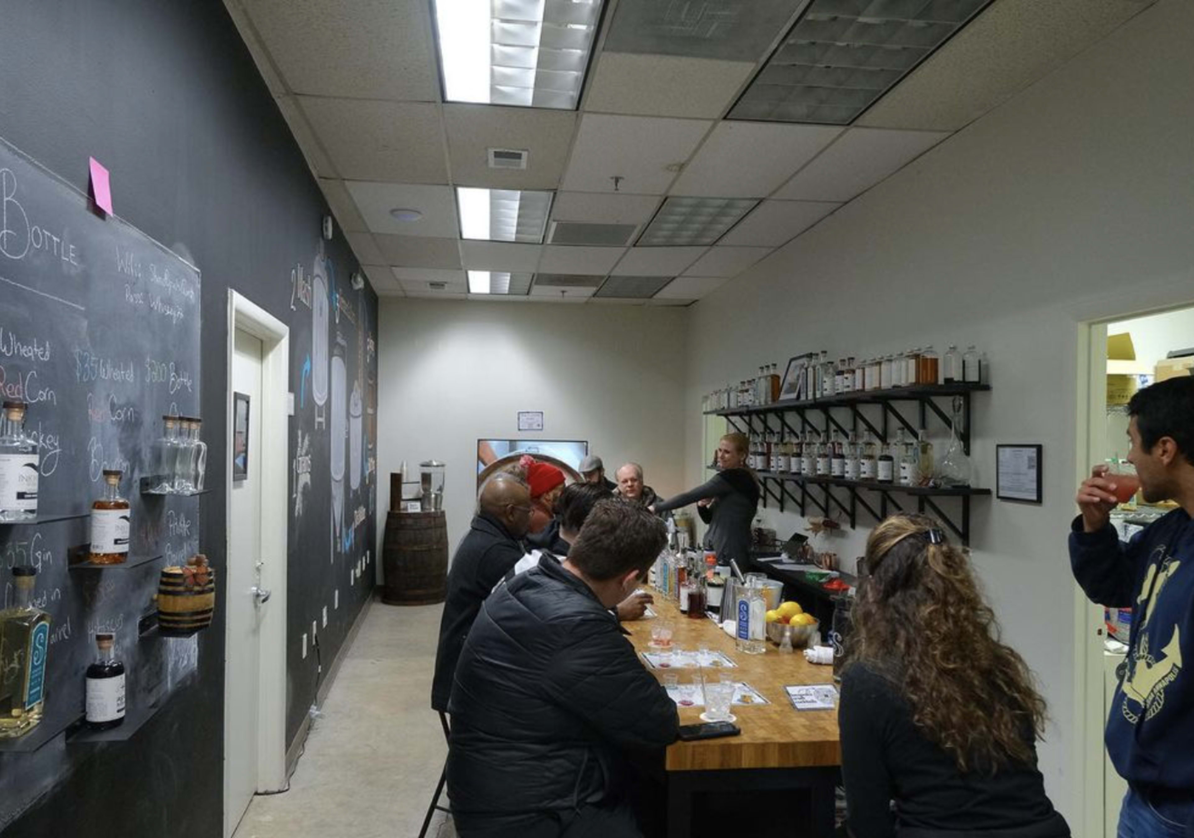 A group of people sits at a long wooden table in a brightly lit room with shelves of bottles and a black chalkboard wall.