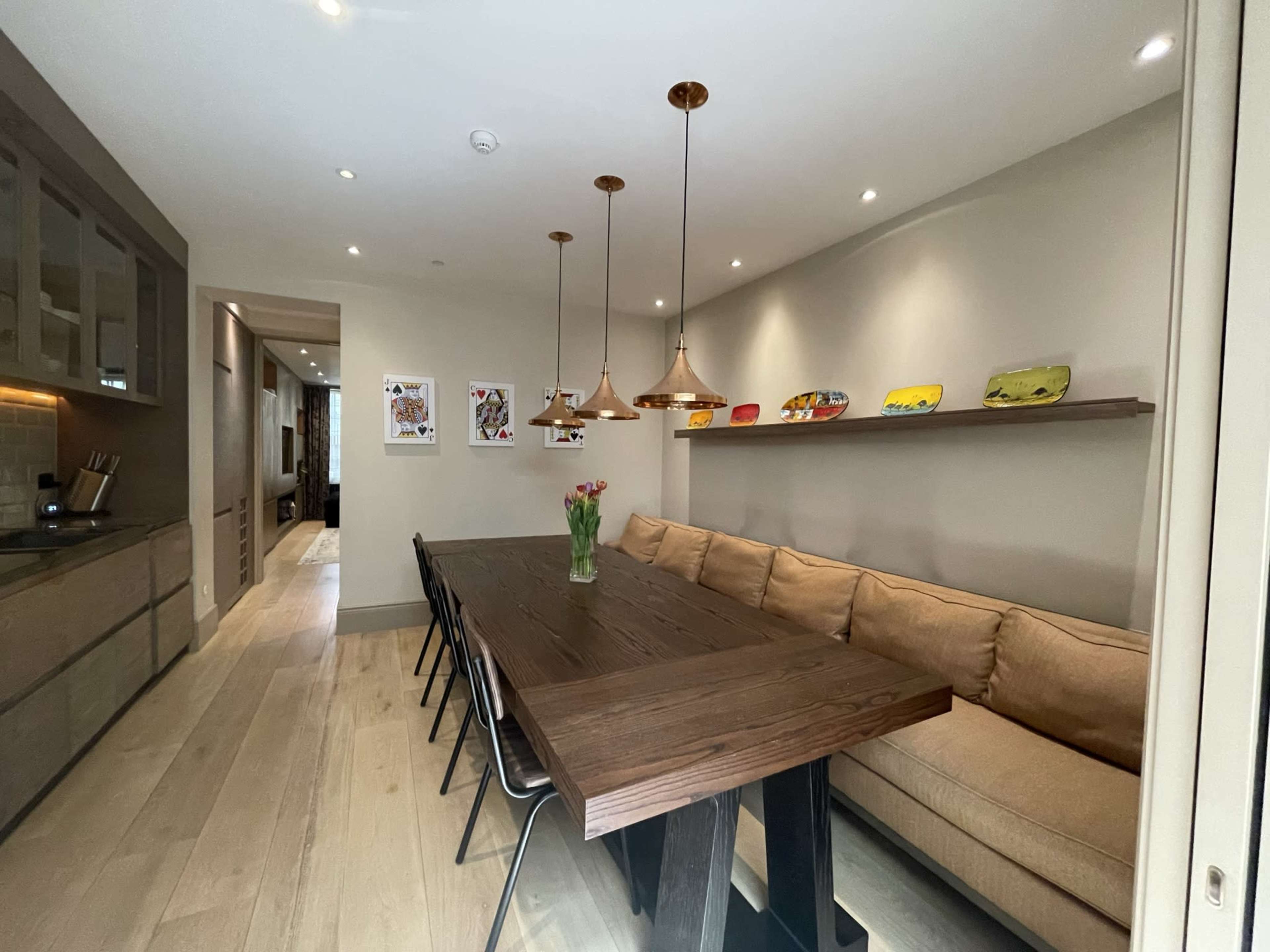 A long wooden dining table with black chairs is positioned in a well-lit room featuring pendant lights and a beige couch along one wall.