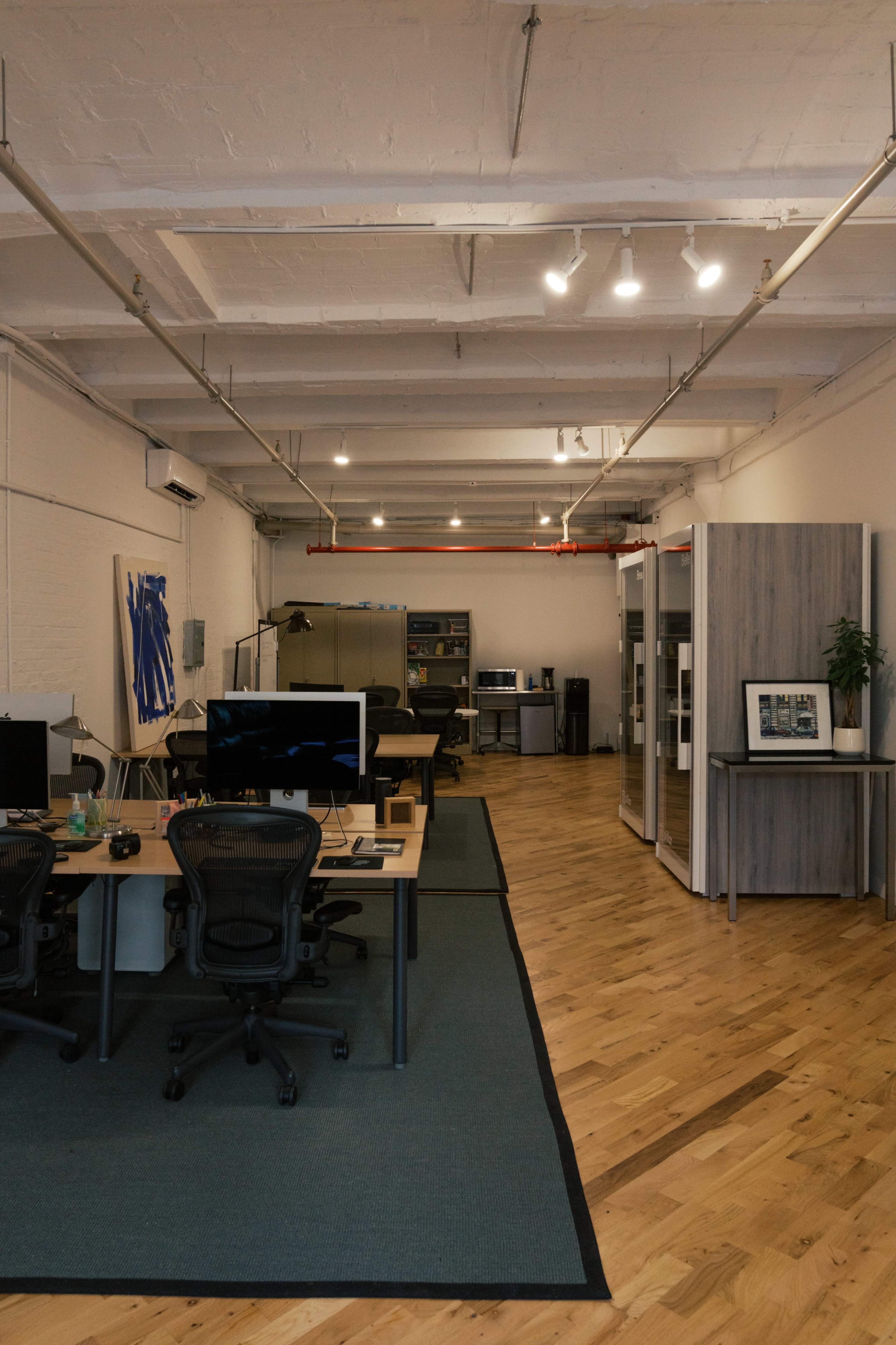 The image shows a modern office space featuring several desks, a wooden floor, and a partitioned area with a plant and framed photo.