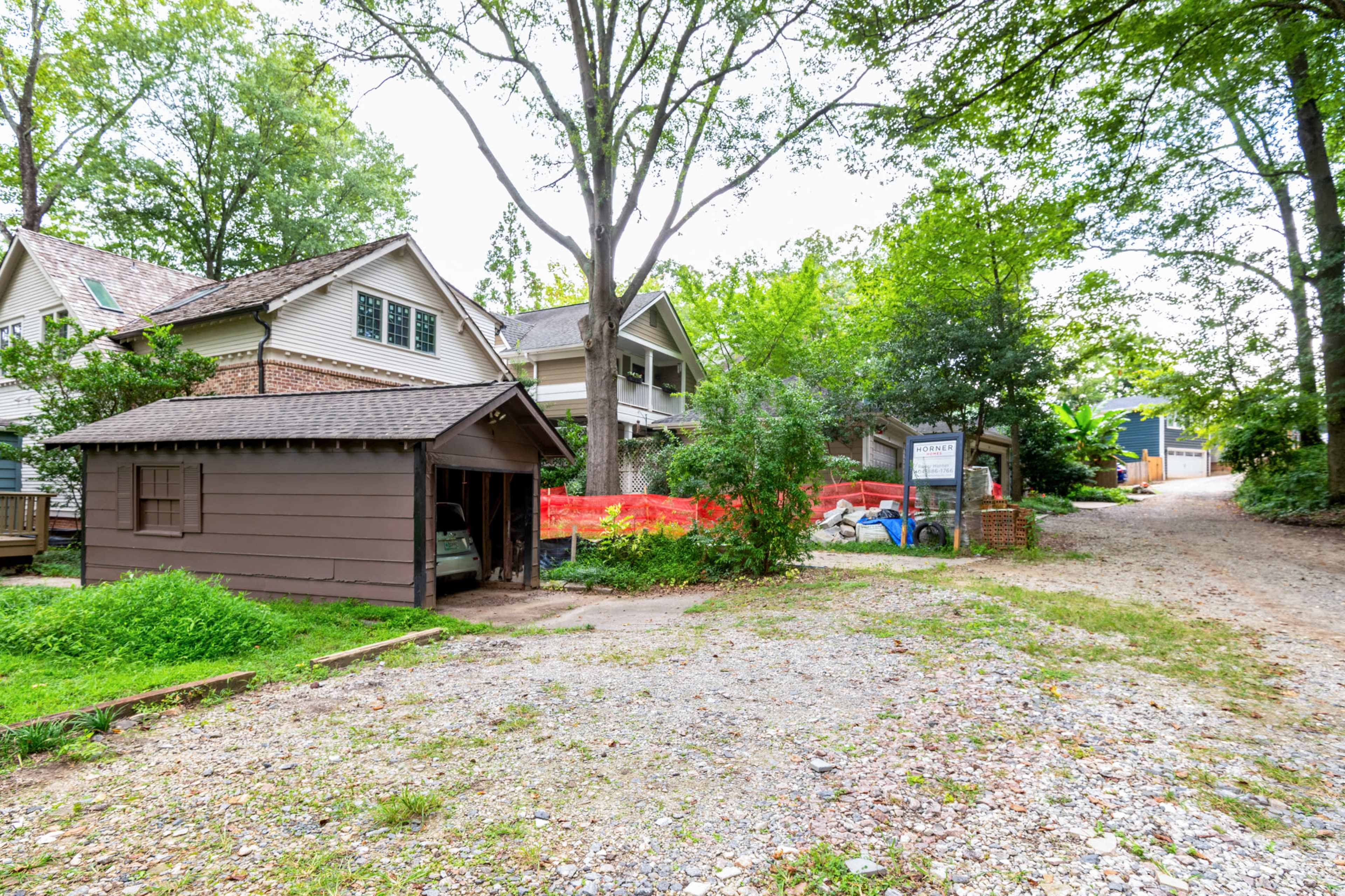 The image shows a gravel path leading away from a brown shed, flanked by trees and houses, with construction barriers visible in the background.