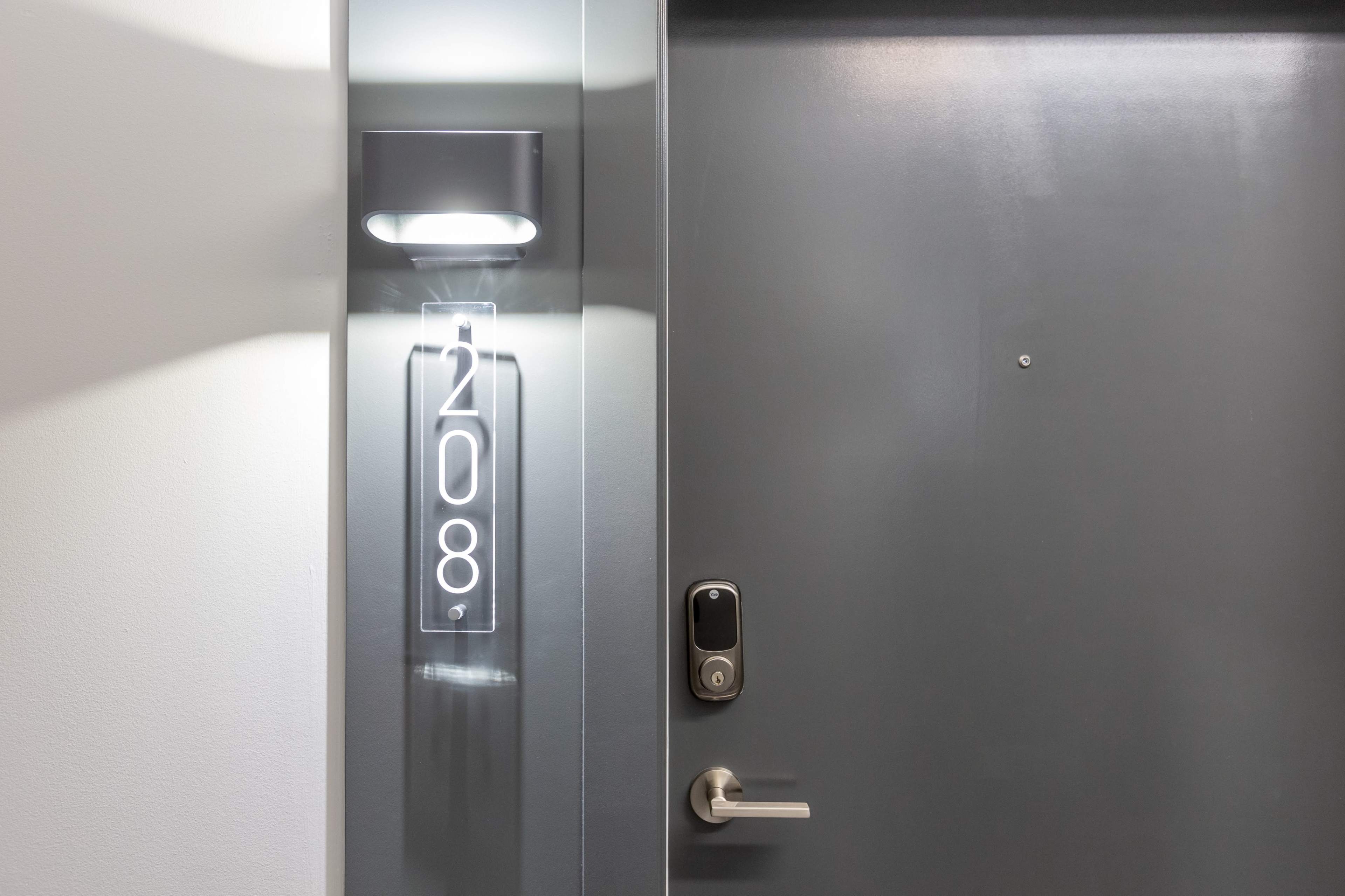 The image shows a modern hotel room door numbered 208, featuring a sleek light fixture and a digital lock.