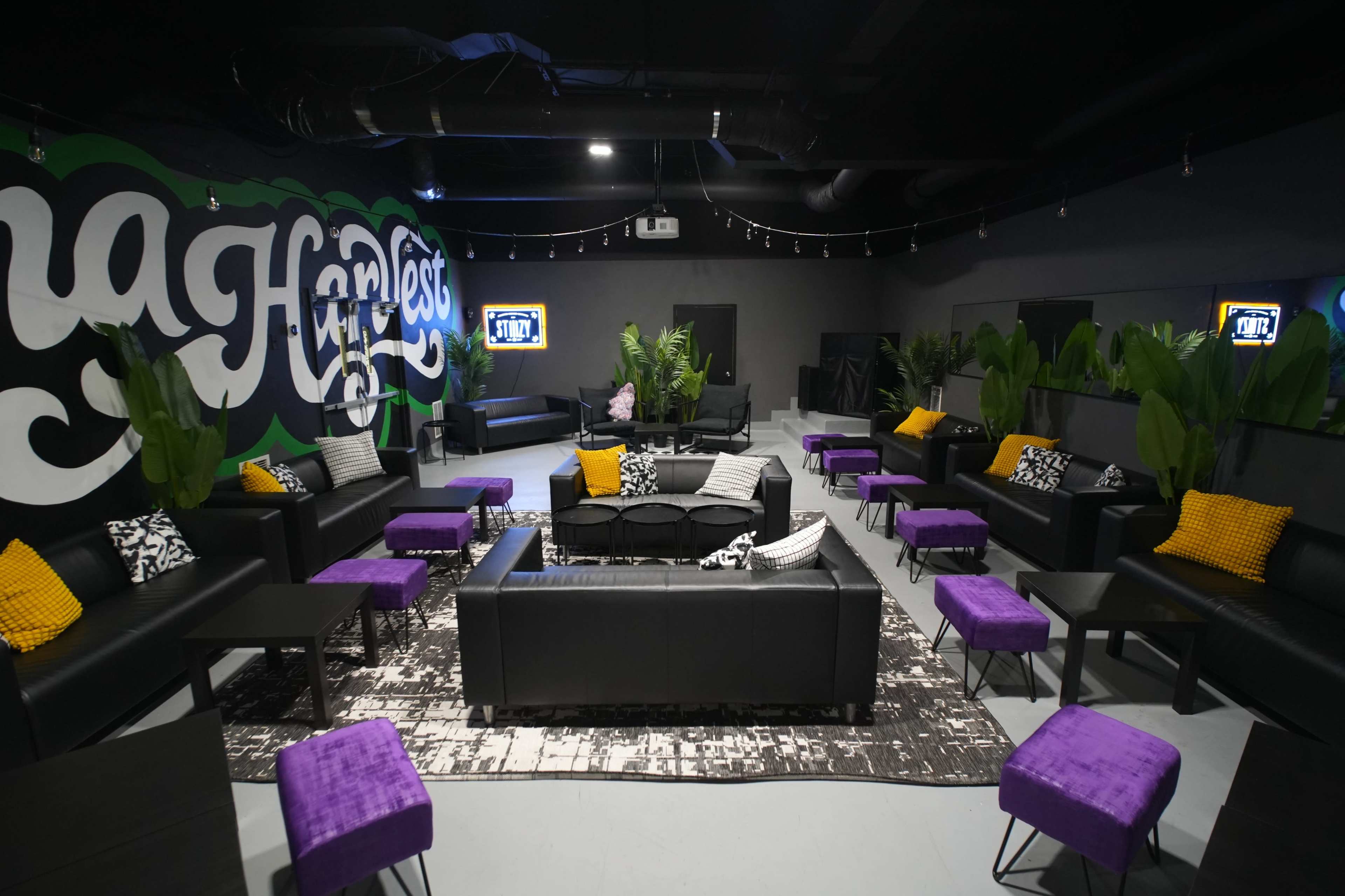 The image shows a modern lounge area with black leather sofas, vibrant purple stools, and a large mural on the wall.