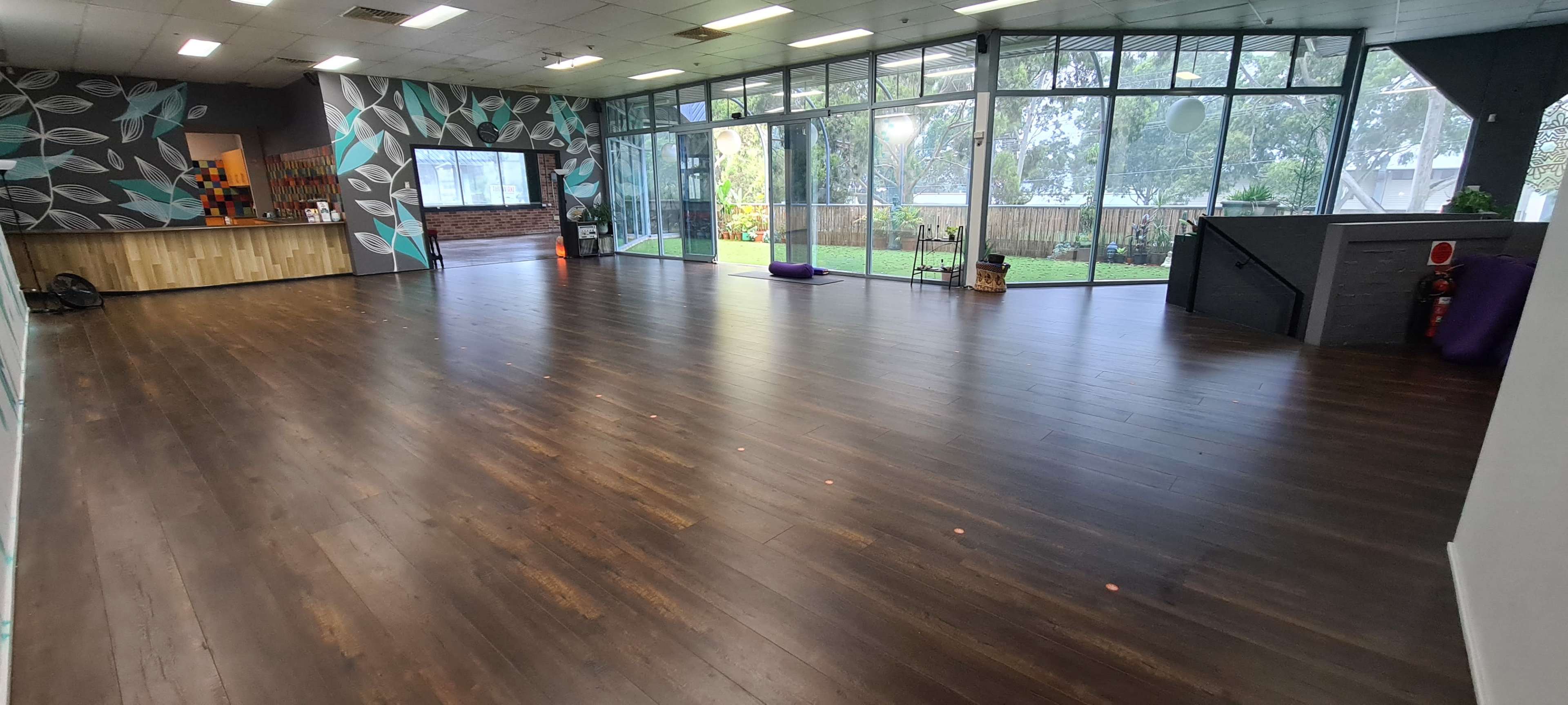 The image shows a spacious indoor area with wooden flooring, large glass windows letting in natural light, and a decorated wall featuring a leaf pattern.