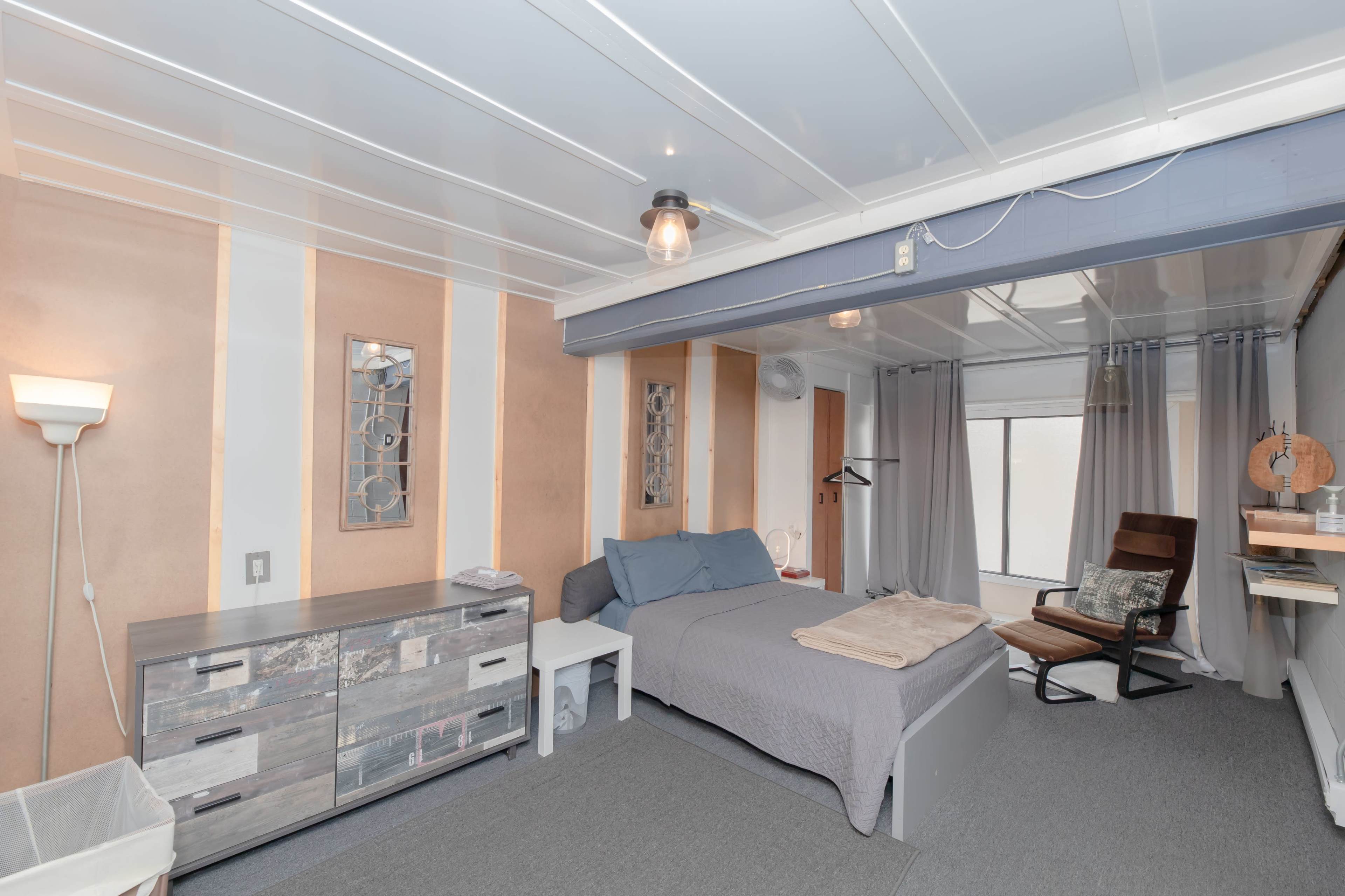 The image shows a contemporary bedroom with a bed, a wooden dresser, a modern chair, and large windows covered by sheer curtains.