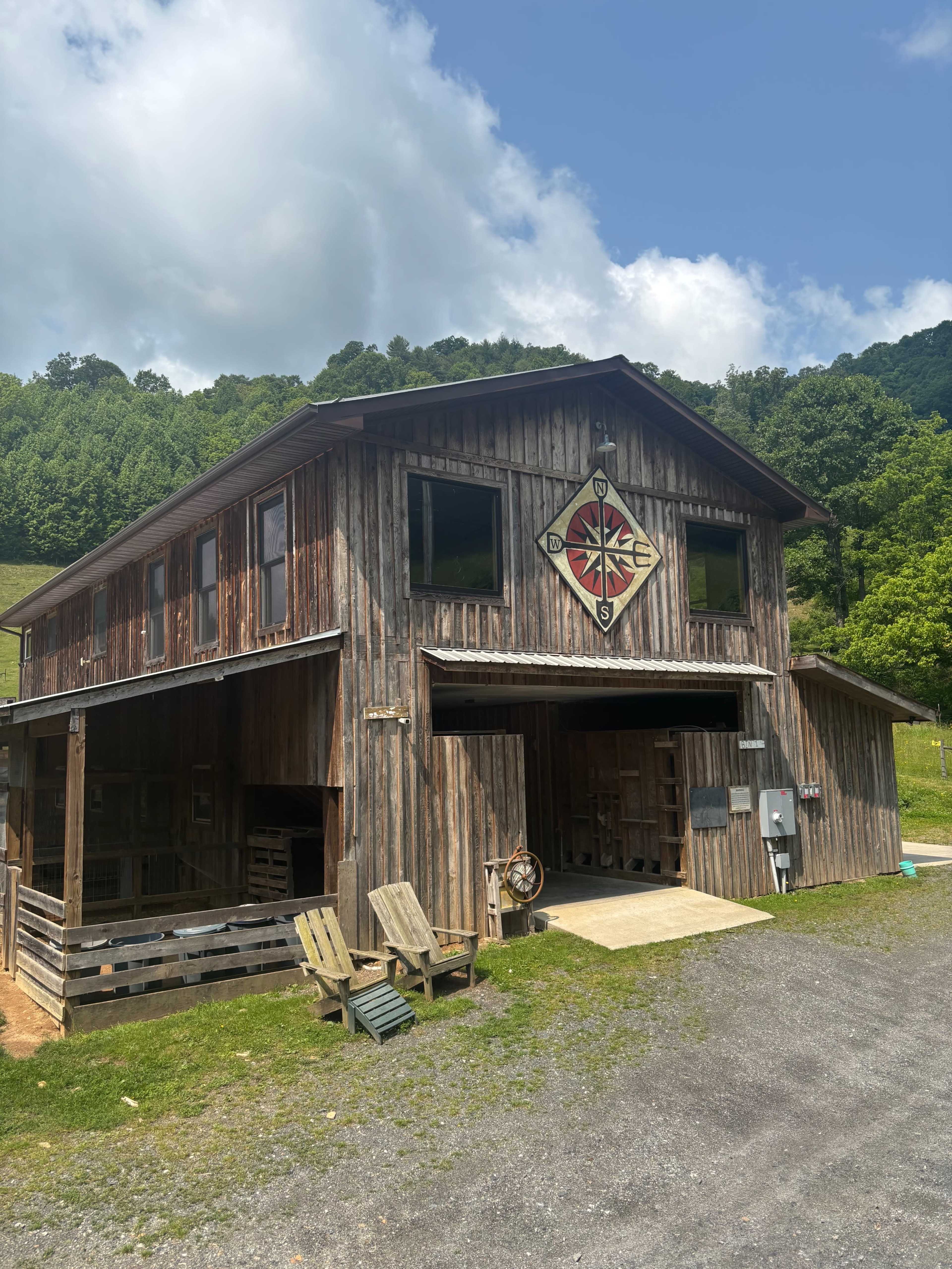A large wooden barn with a compass rose emblem on the front is situated in a green, hilly landscape under a partly cloudy sky.