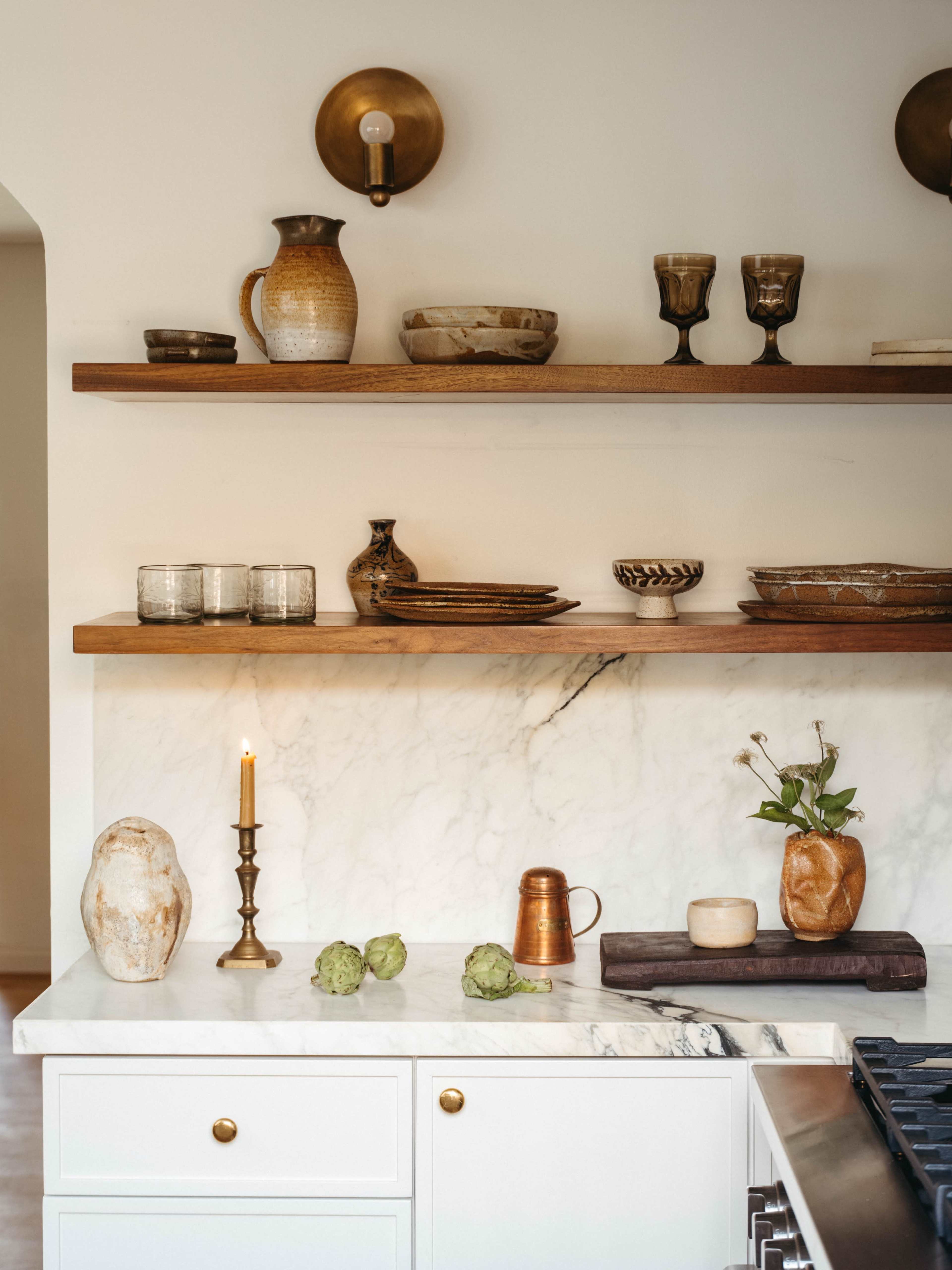The image features a kitchen countertop with marble surface, displaying ceramic vessels, glassware, and a candle holder, all arranged on wooden shelves.
