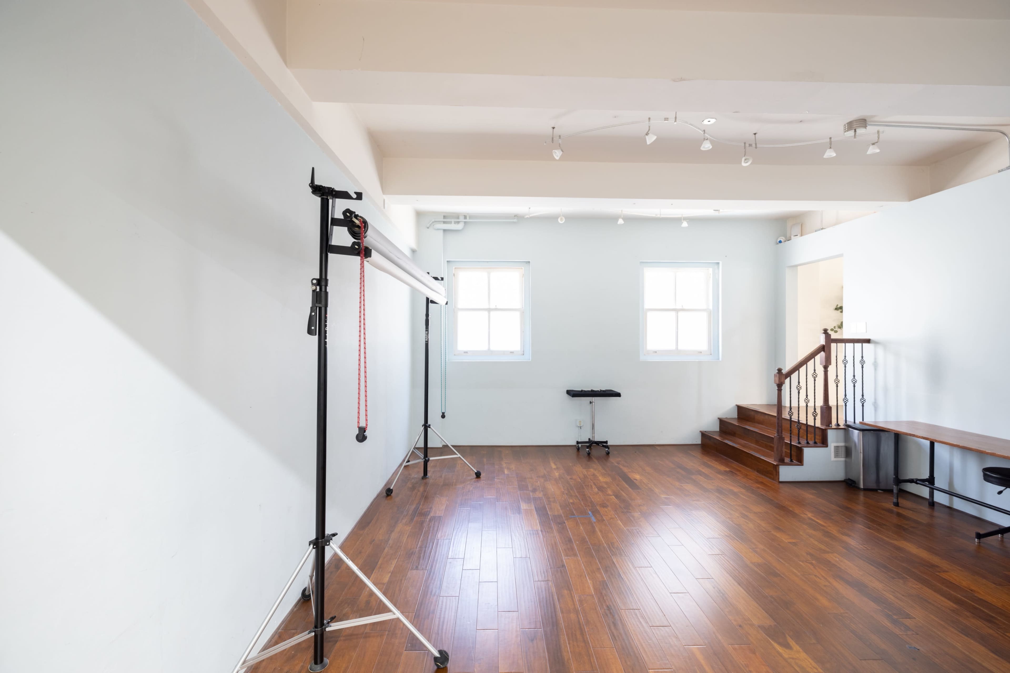 The image shows a spacious, empty room with wood flooring, a backdrop setup, a bench, and a staircase.