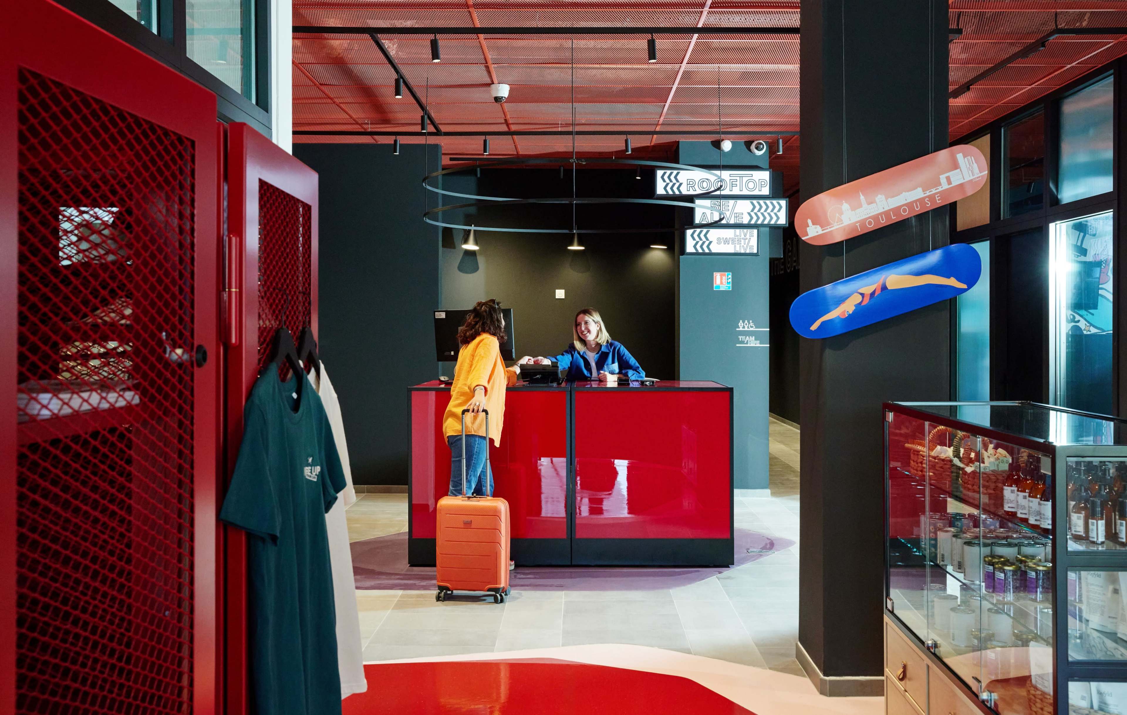 A traveler with a suitcase is checking in at a red reception desk while a staff member assists them in a modern lobby.