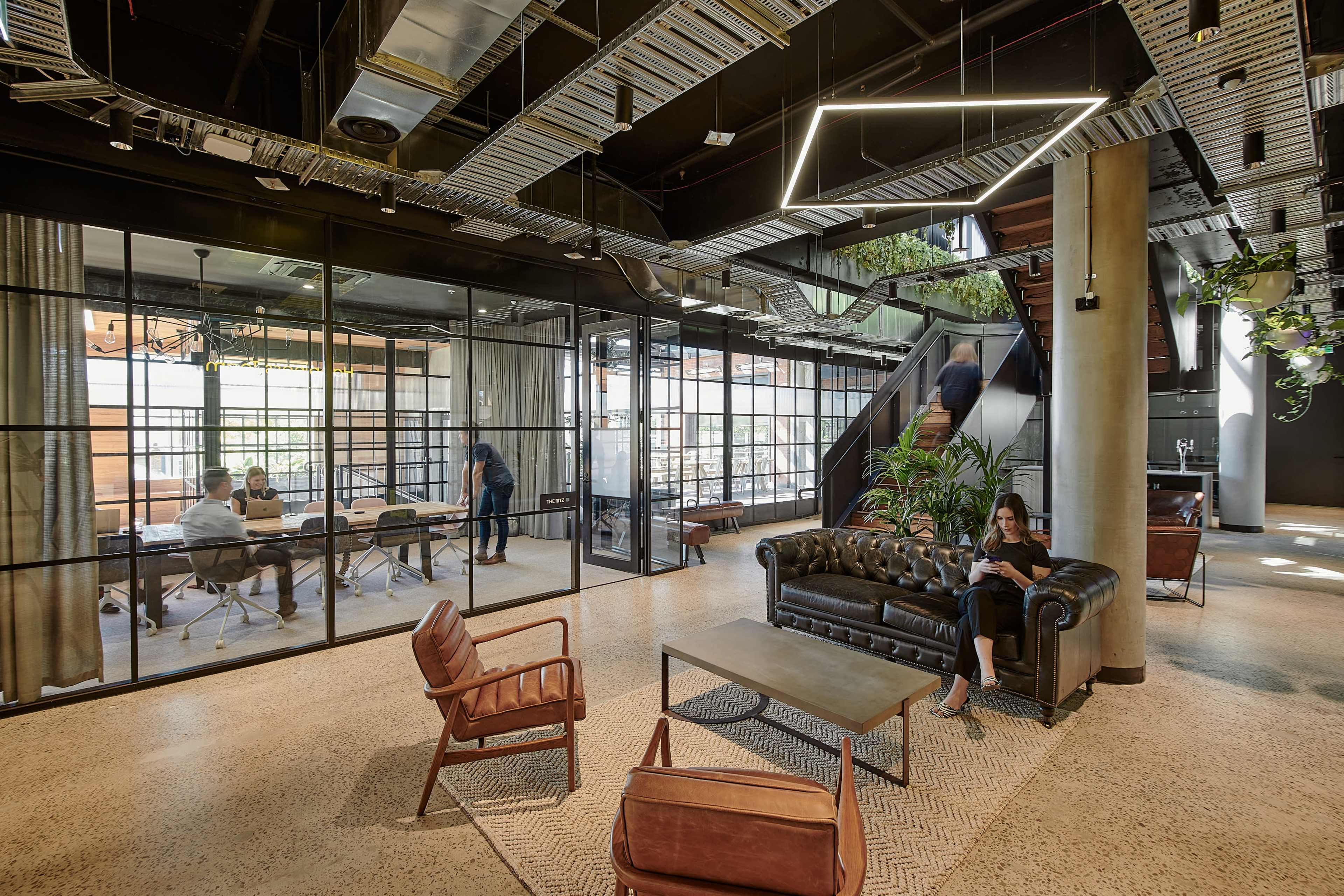 The image shows a modern office space featuring a lounge area with a leather sofa and armchairs, while glass-walled meeting rooms and a staircase are visible in the background.