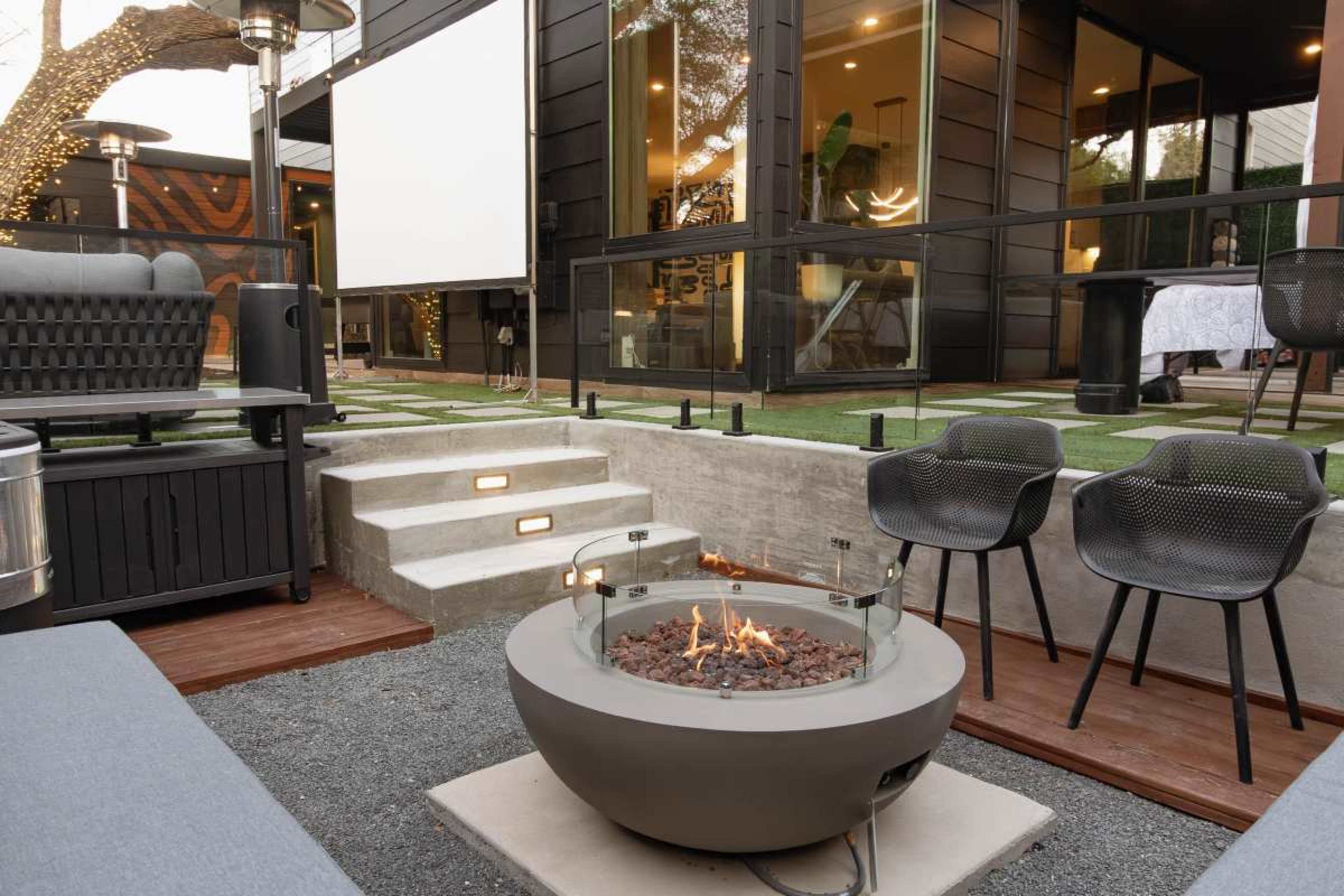 The image shows a modern outdoor seating area featuring a circular fire pit with flames, surrounded by minimalistic chairs and steps leading to a glass-walled building.