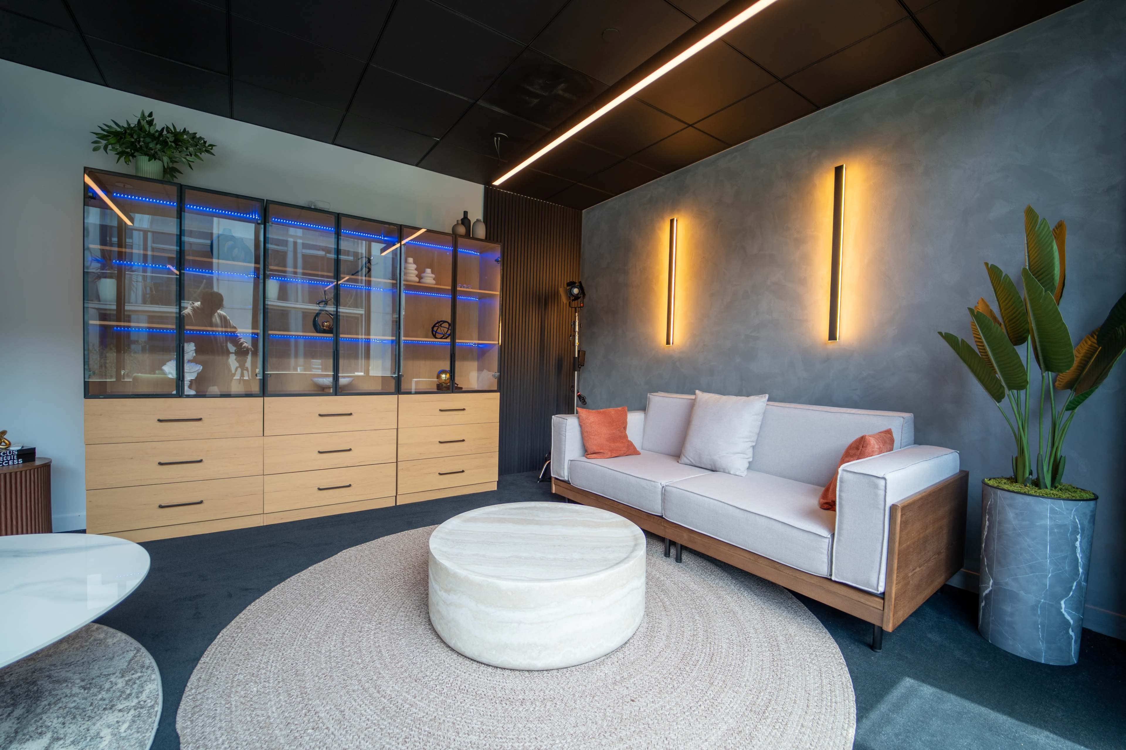 The image shows a modern lounge area featuring a light gray sofa, a round ottoman, and a wooden storage unit with glass doors illuminated by blue lighting.