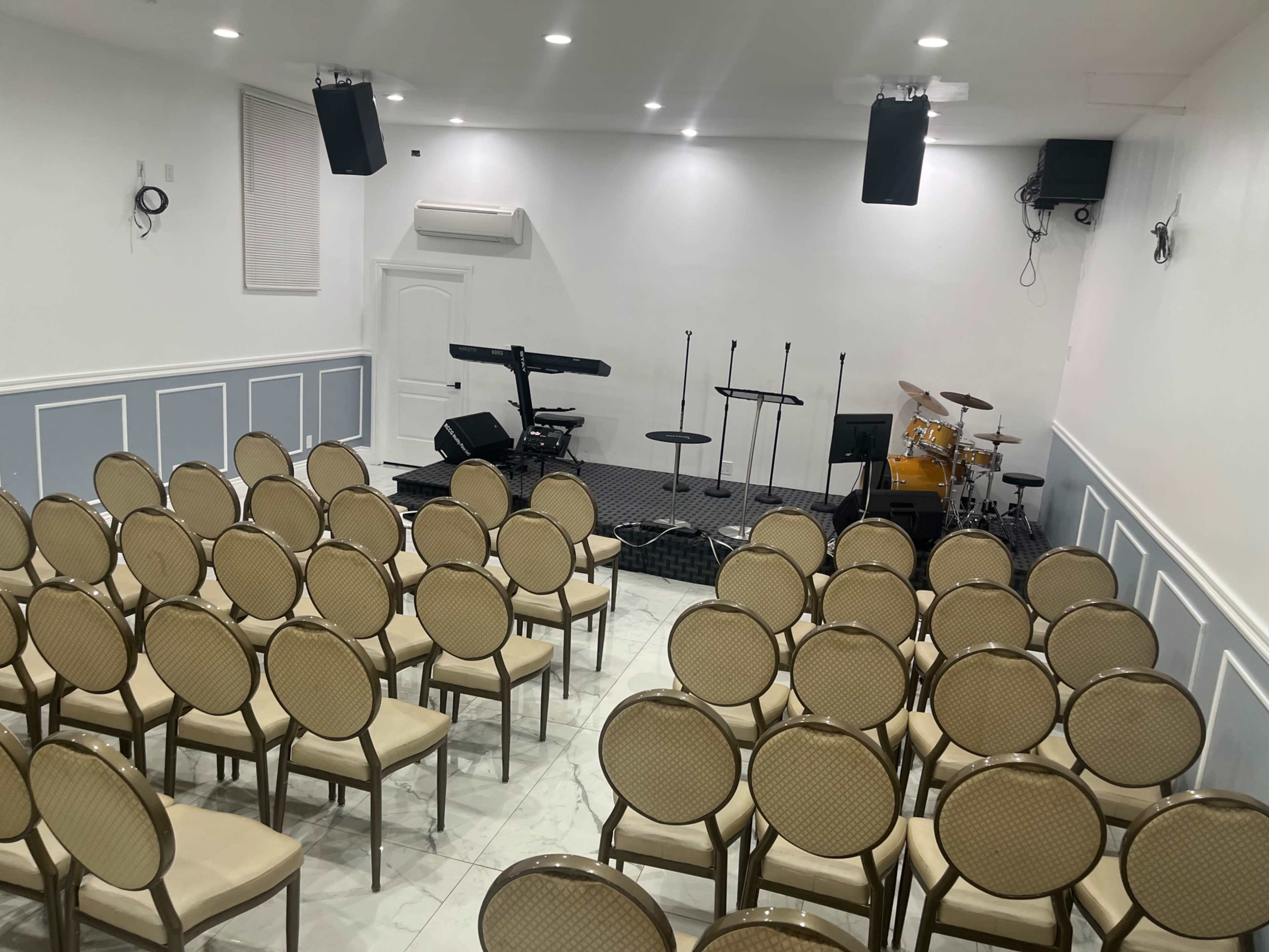 A room set up for a performance, featuring rows of empty chairs facing a stage equipped with musical instruments, including a keyboard and a drum set.
