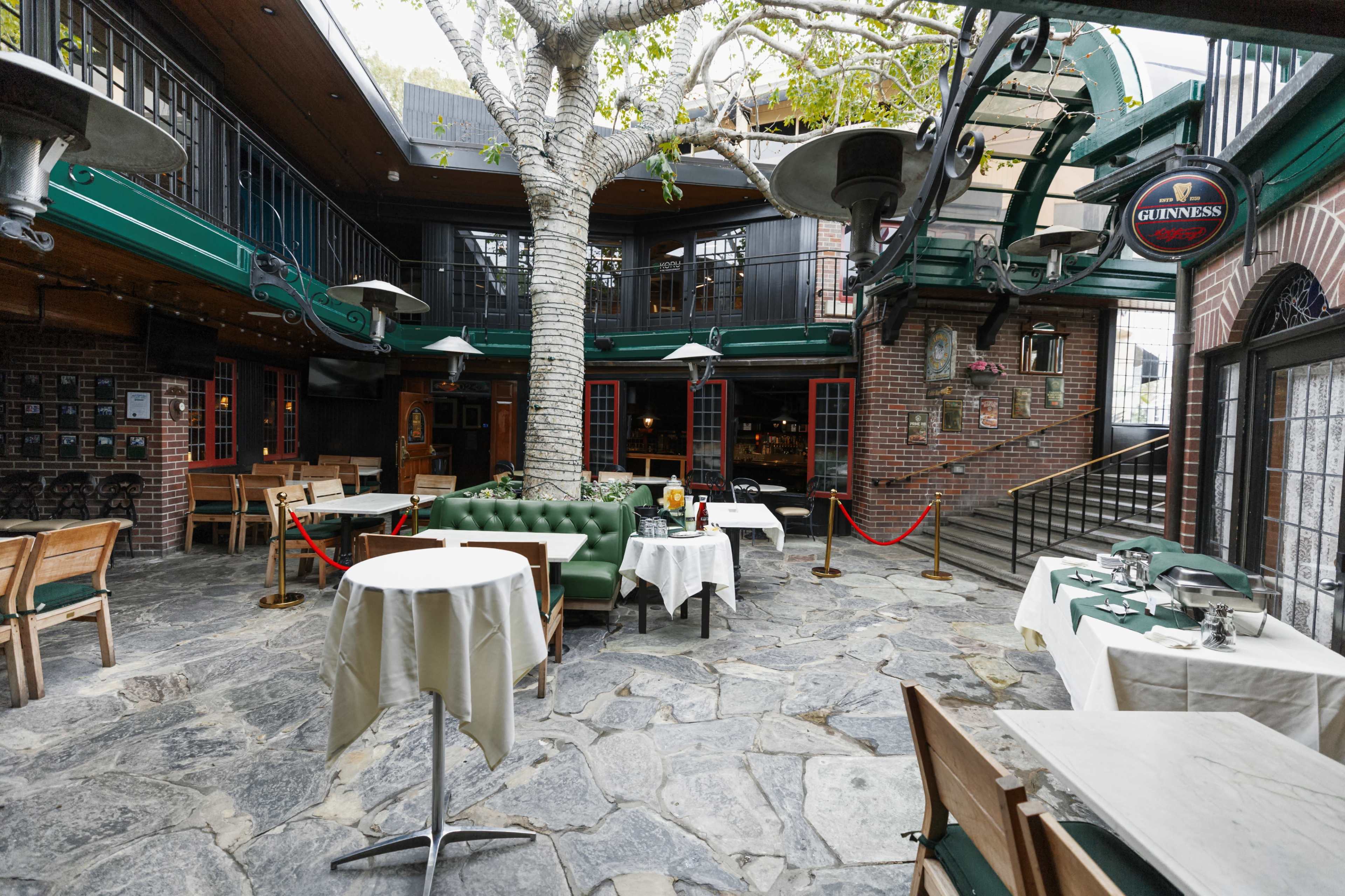 The image shows a spacious, outdoor restaurant area featuring stone flooring, green furnishings, and a large tree in the center.
