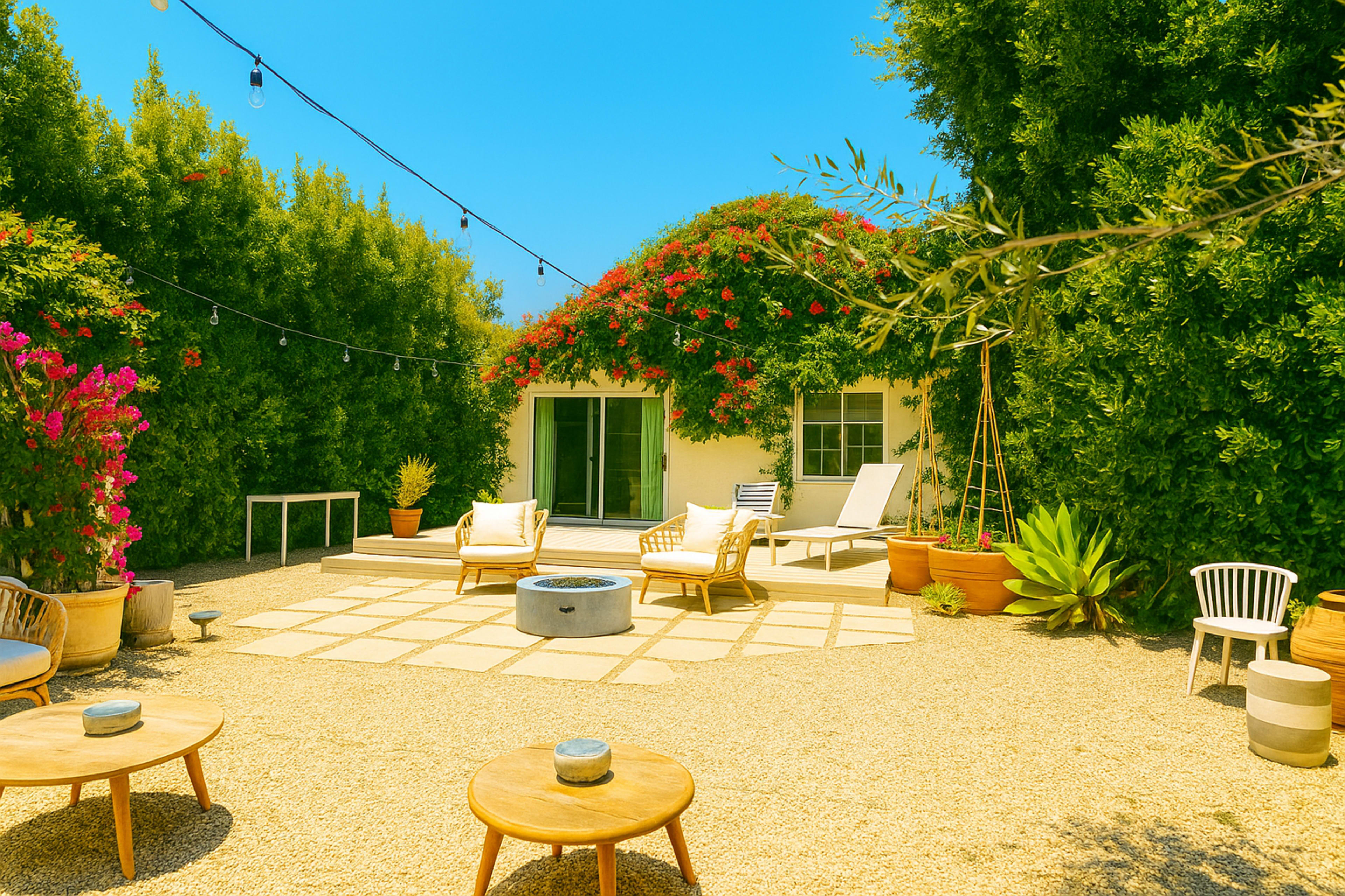 A backyard patio features a stone seating area with a fire pit, surrounded by greenery and decorative plants, along with lounge chairs and hanging string lights above.