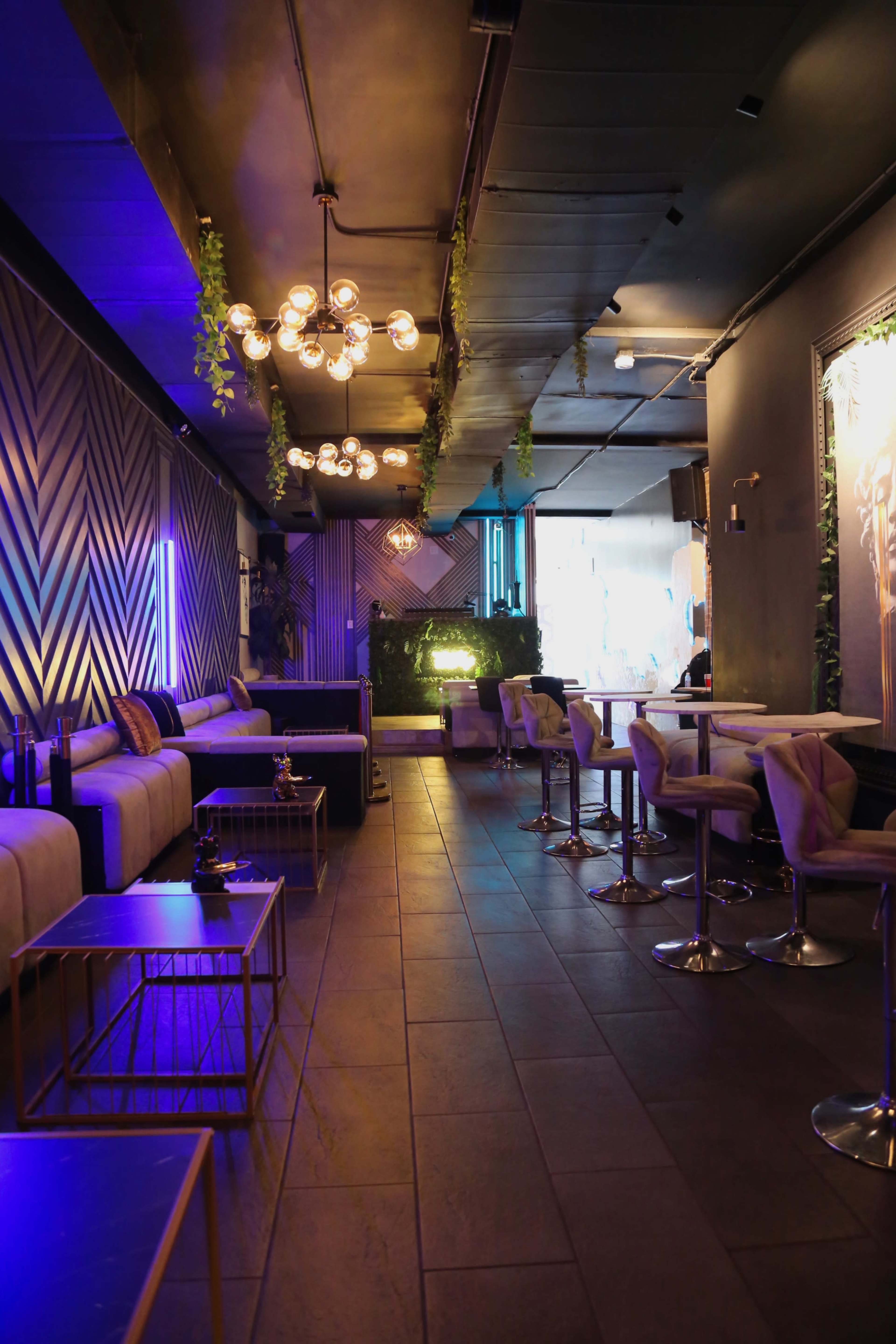 A dimly lit lounge with modern seating, decorative lighting, and plants, leading to an illuminated area at the back.