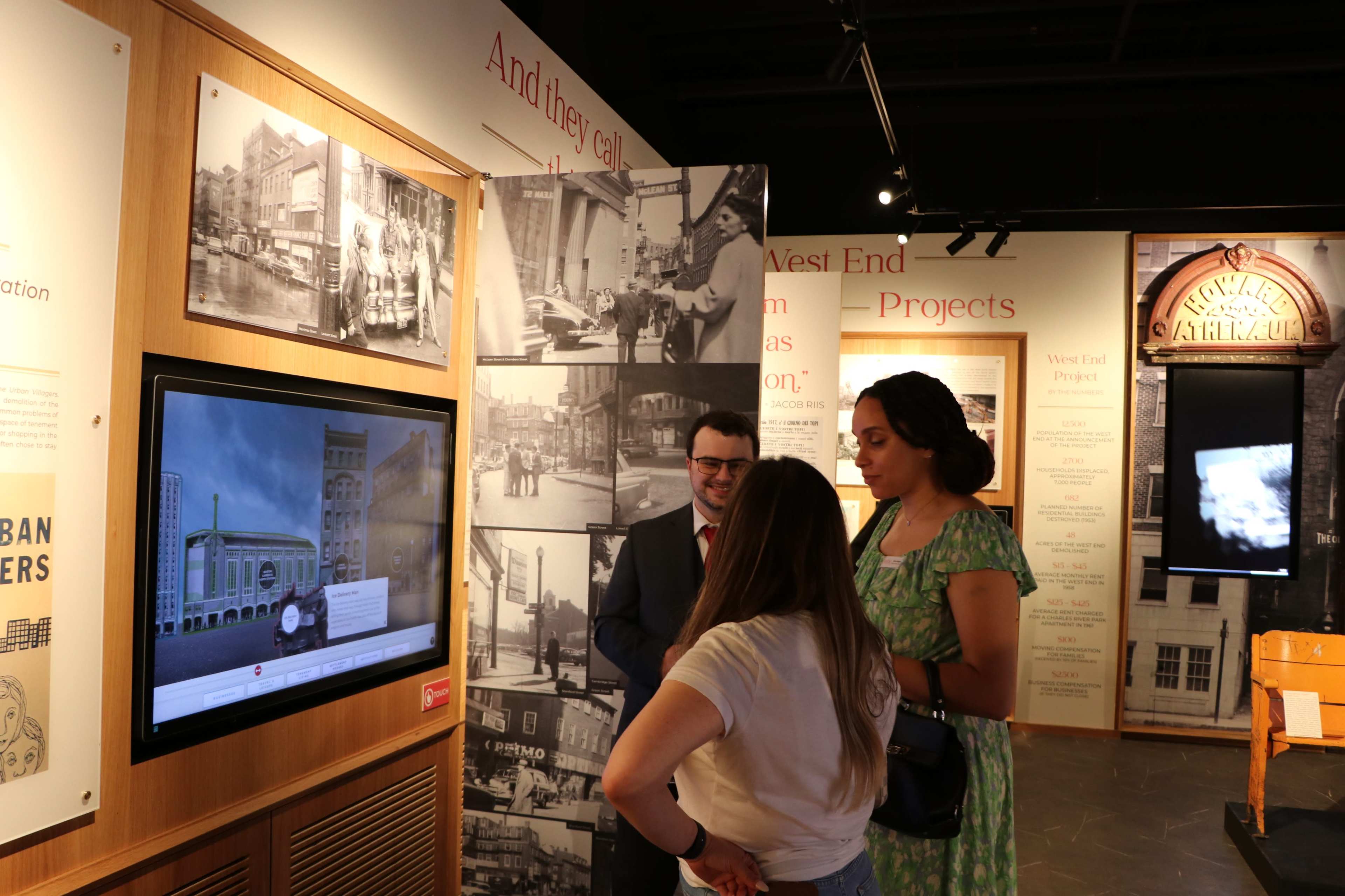 Three visitors engage with an interactive display in a museum, surrounded by historical photographs and information about urban development projects.