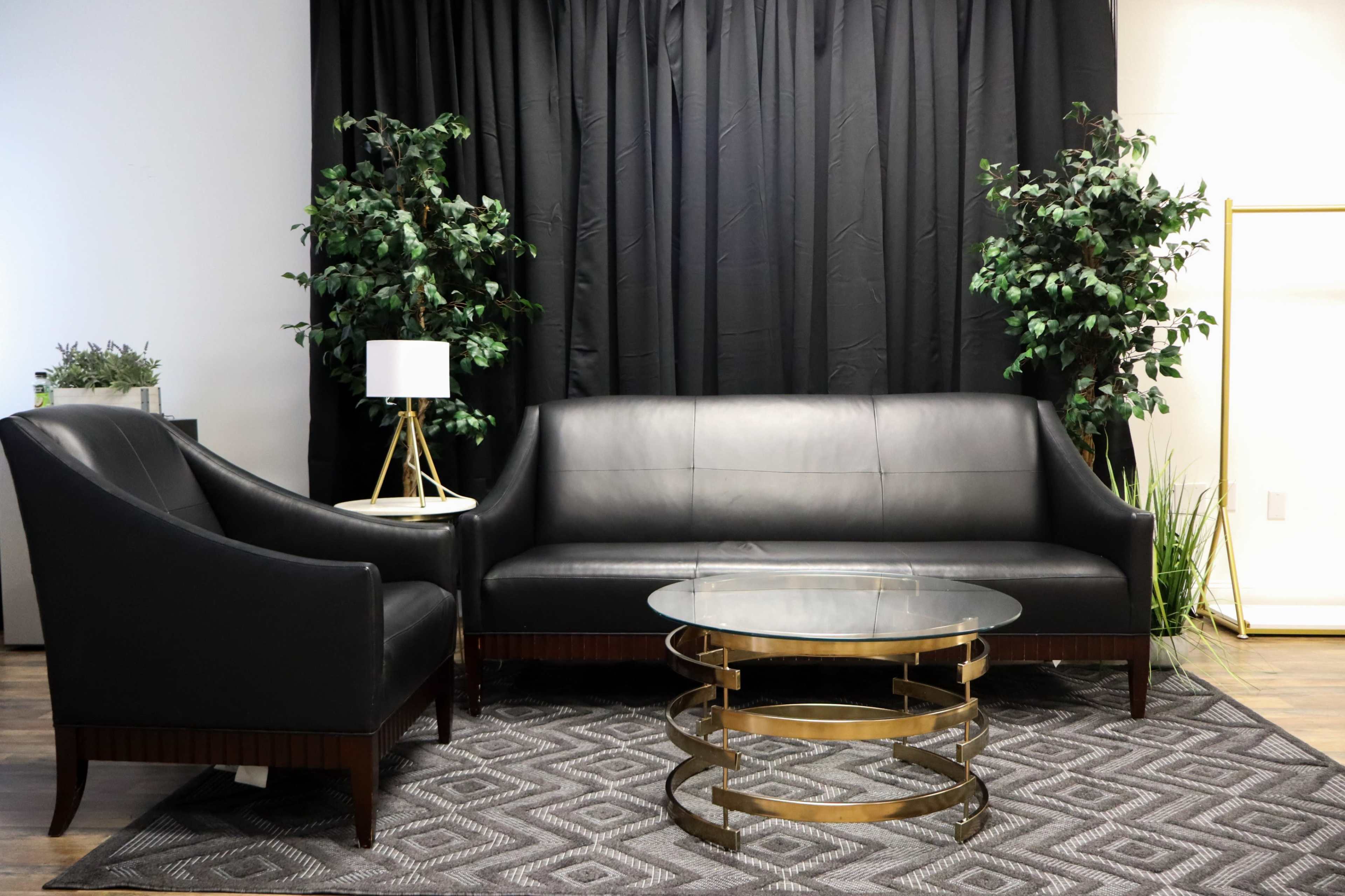 The image shows a modern living room setup featuring a black leather sofa and armchair, a circular glass coffee table with a gold base, and green potted plants against a black curtain backdrop.