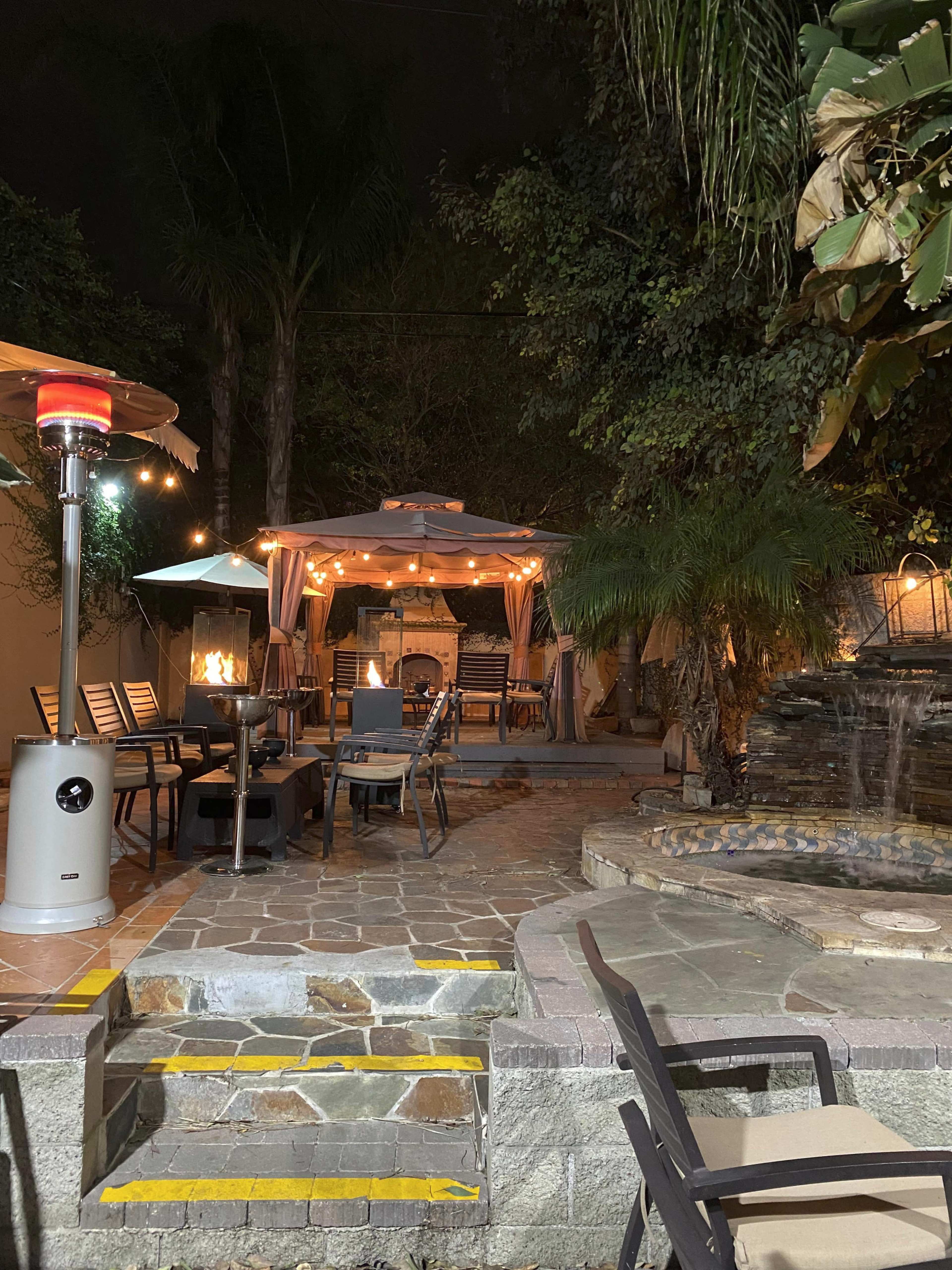 A well-lit outdoor patio with a gazebo, a fire pit, seating arrangements, and a small water feature surrounded by lush greenery.