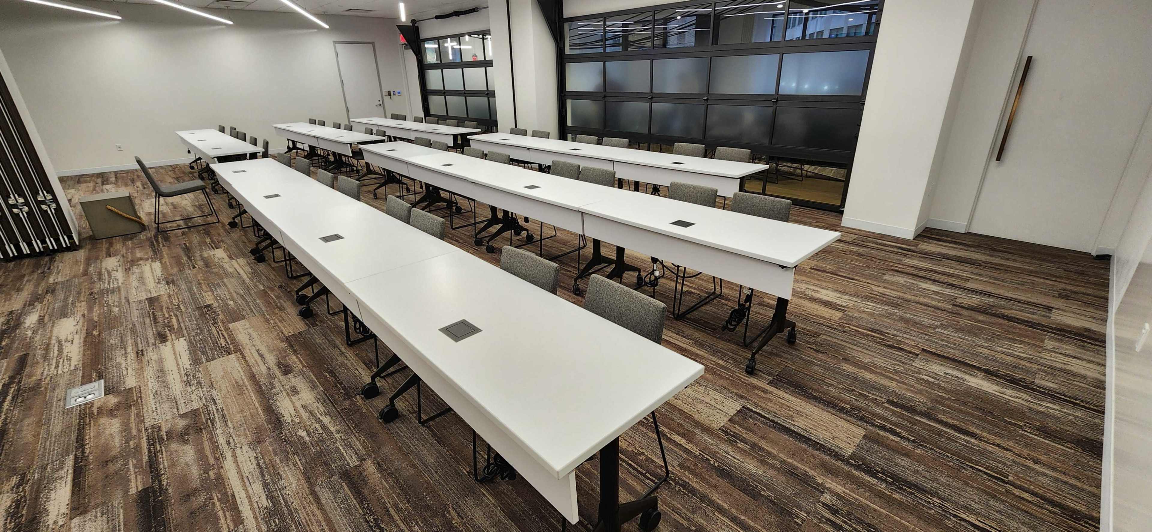A spacious, modern classroom arranged with multiple rows of white tables and gray chairs on a wooden floor.