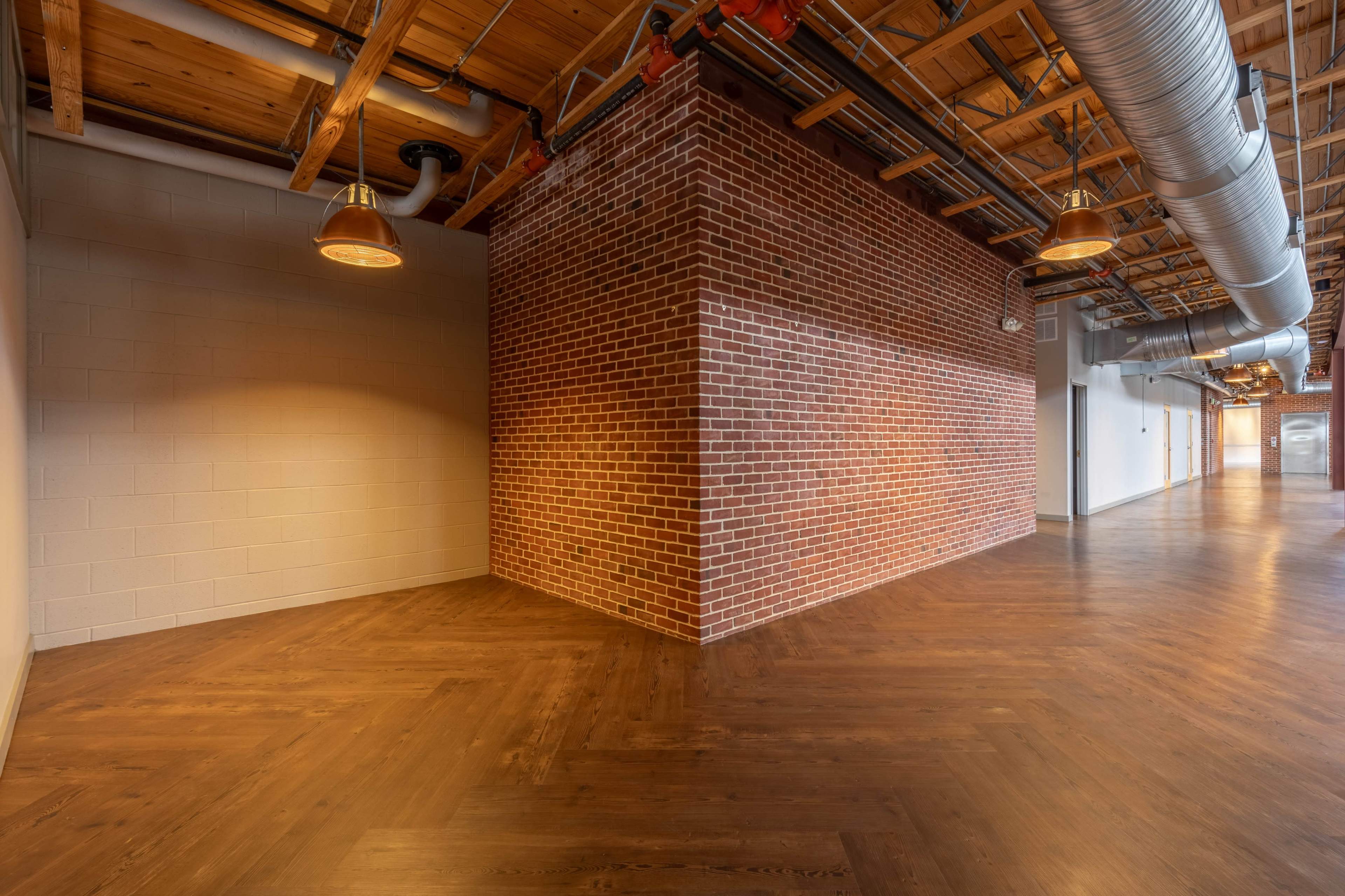 The image shows a corridor with a wooden floor and a brick wall at the corner, accompanied by metal ductwork and overhead lights.