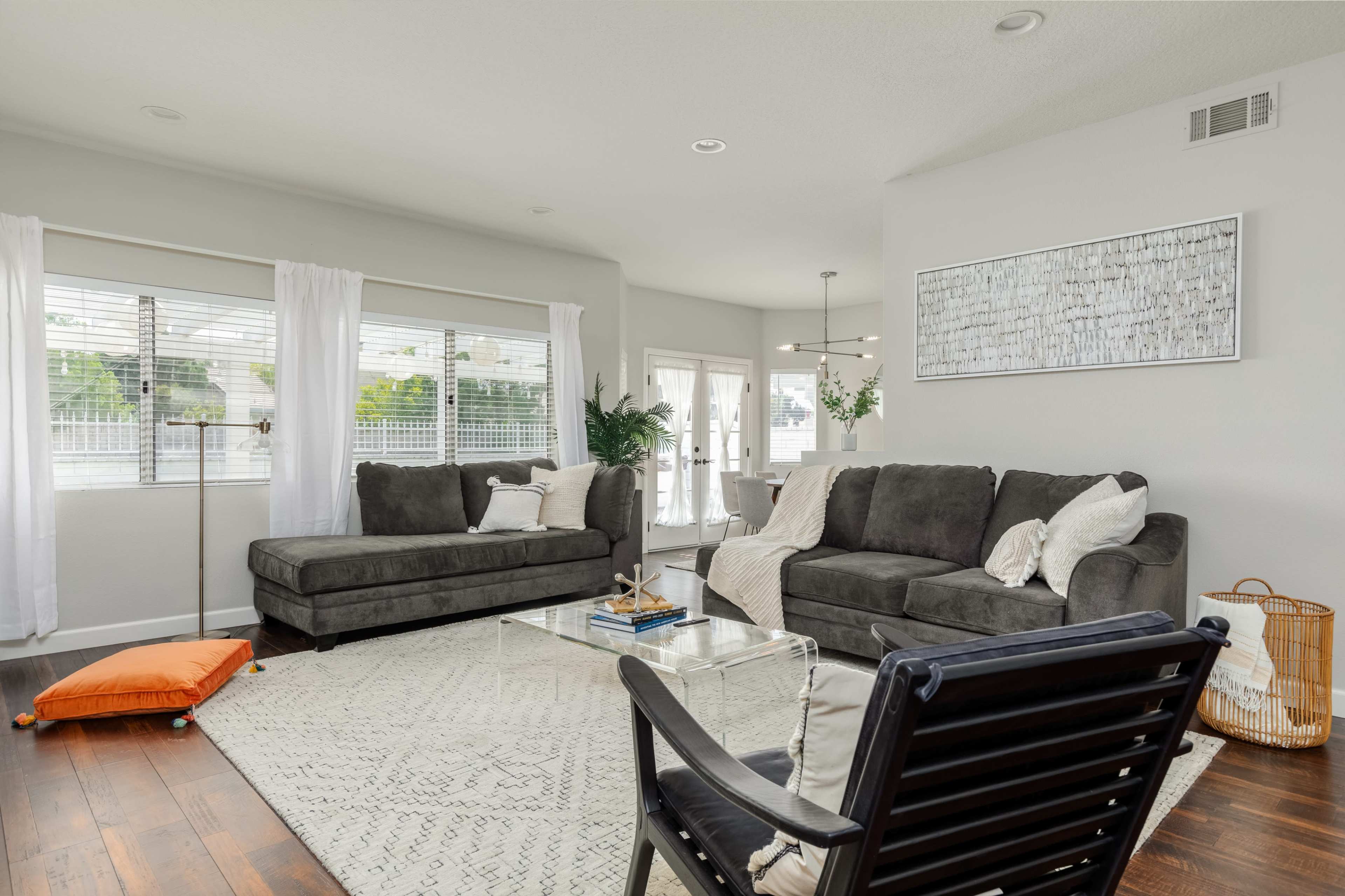 The image shows a modern living room with a dark sectional sofa, a glass coffee table, and a patterned area rug, illuminated by natural light from large windows.