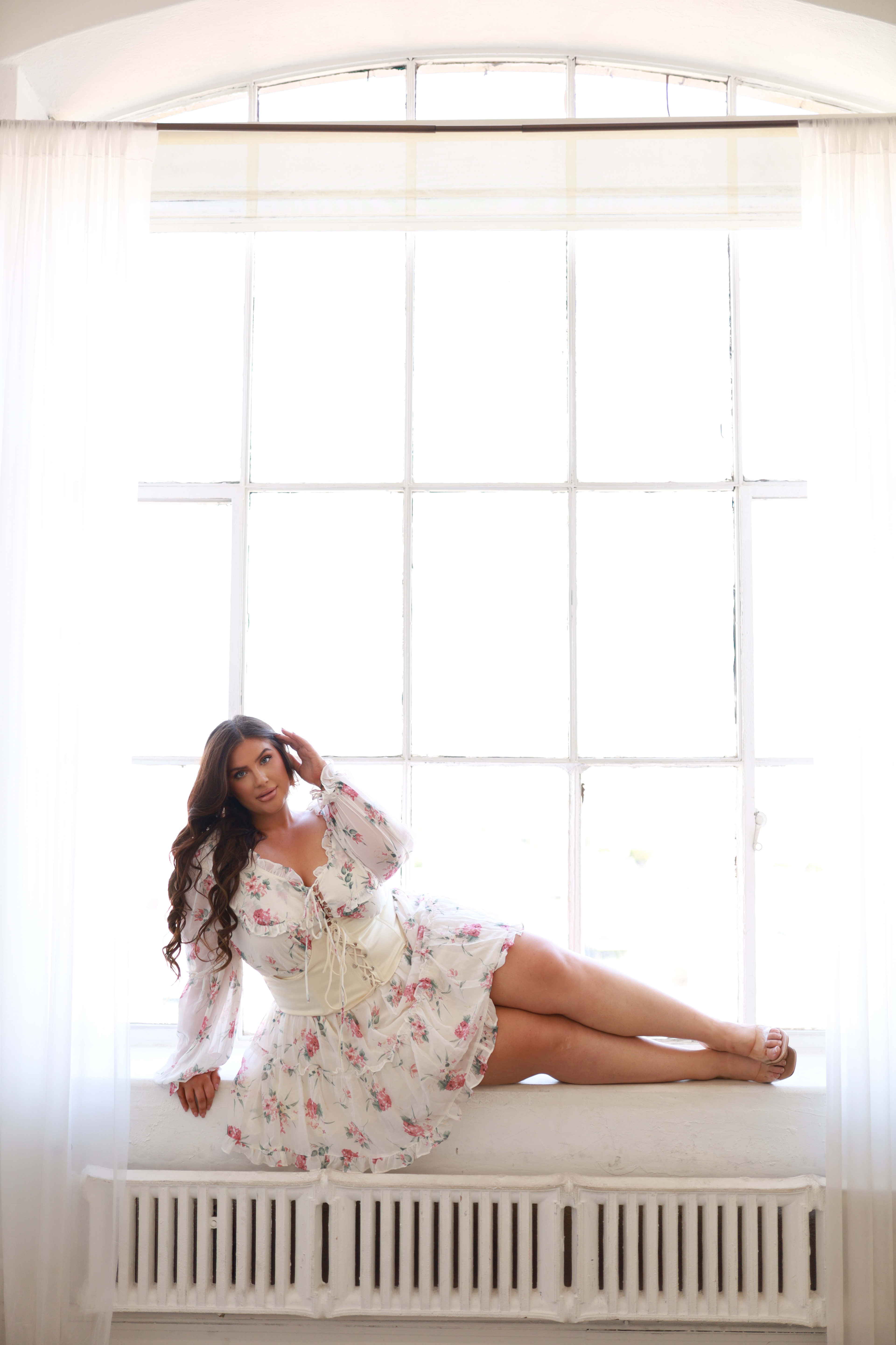 A woman posing for a renaissance fashion photoshoot in a white dress while sitting on a window sill.
