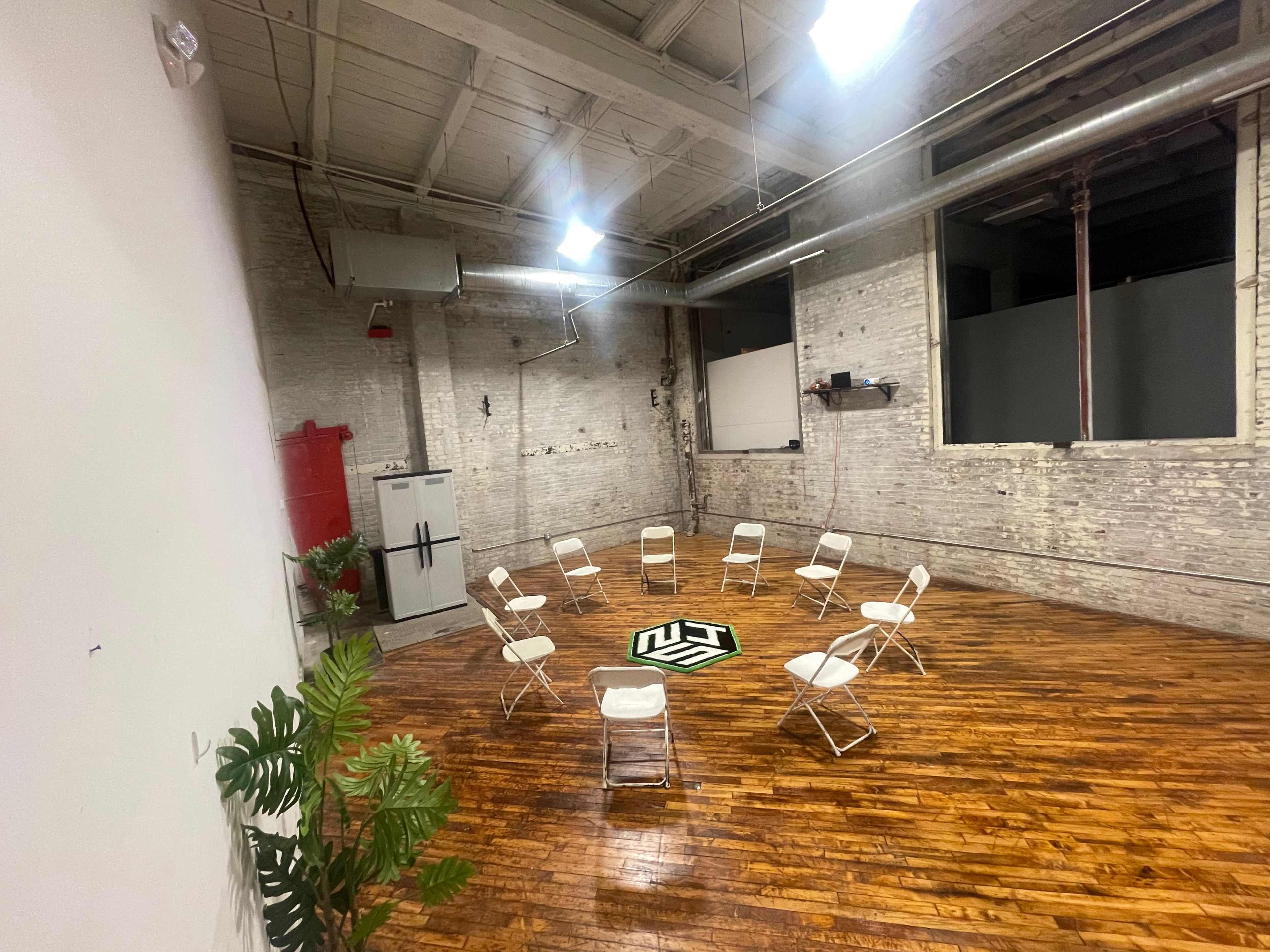 The image shows an empty room with a wooden floor, featuring a circle of white folding chairs arranged around a central symbol on the floor.