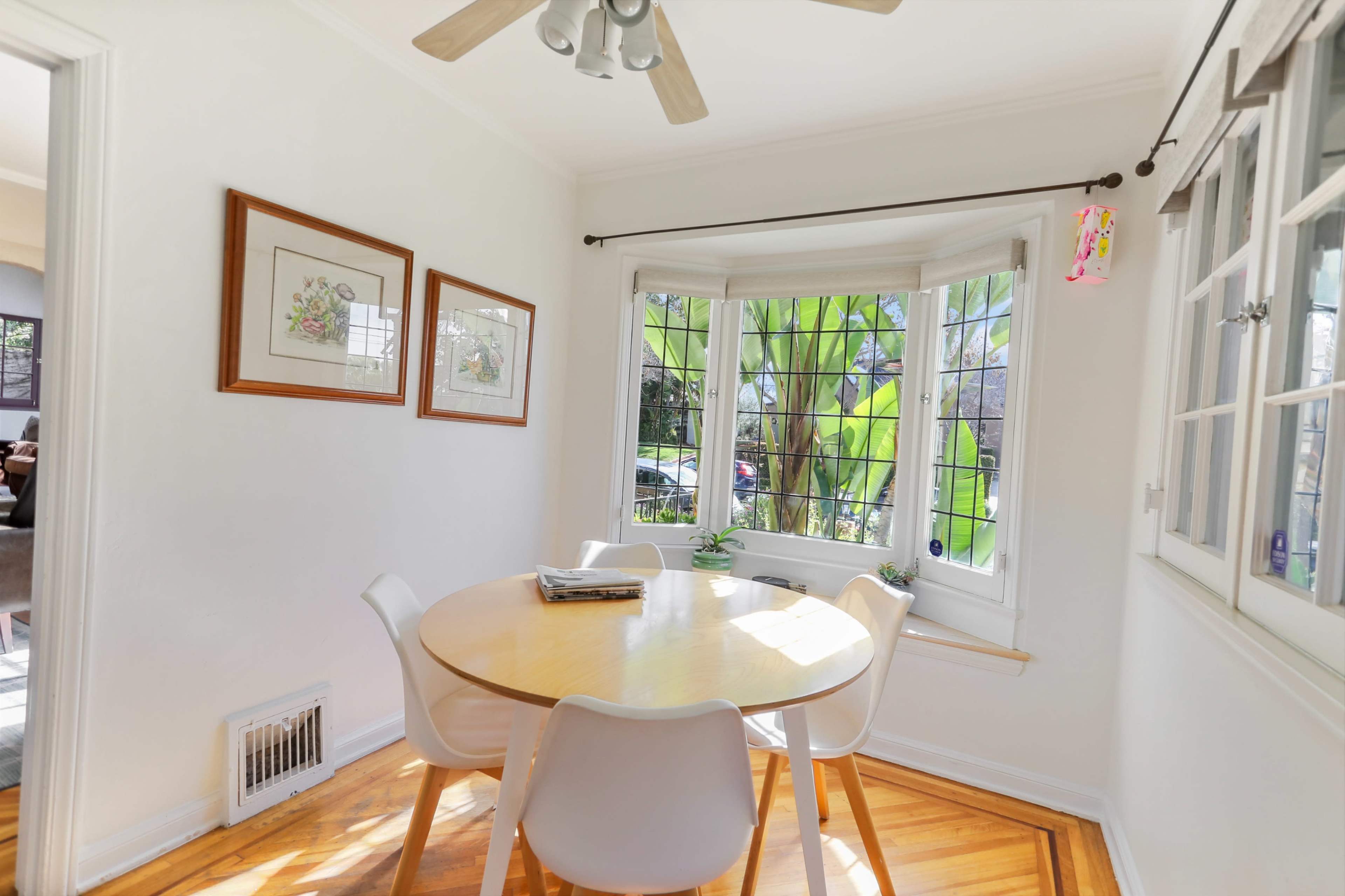 A round wooden dining table with four white chairs is positioned near a large bay window adorned with plants and framed artwork on the walls.