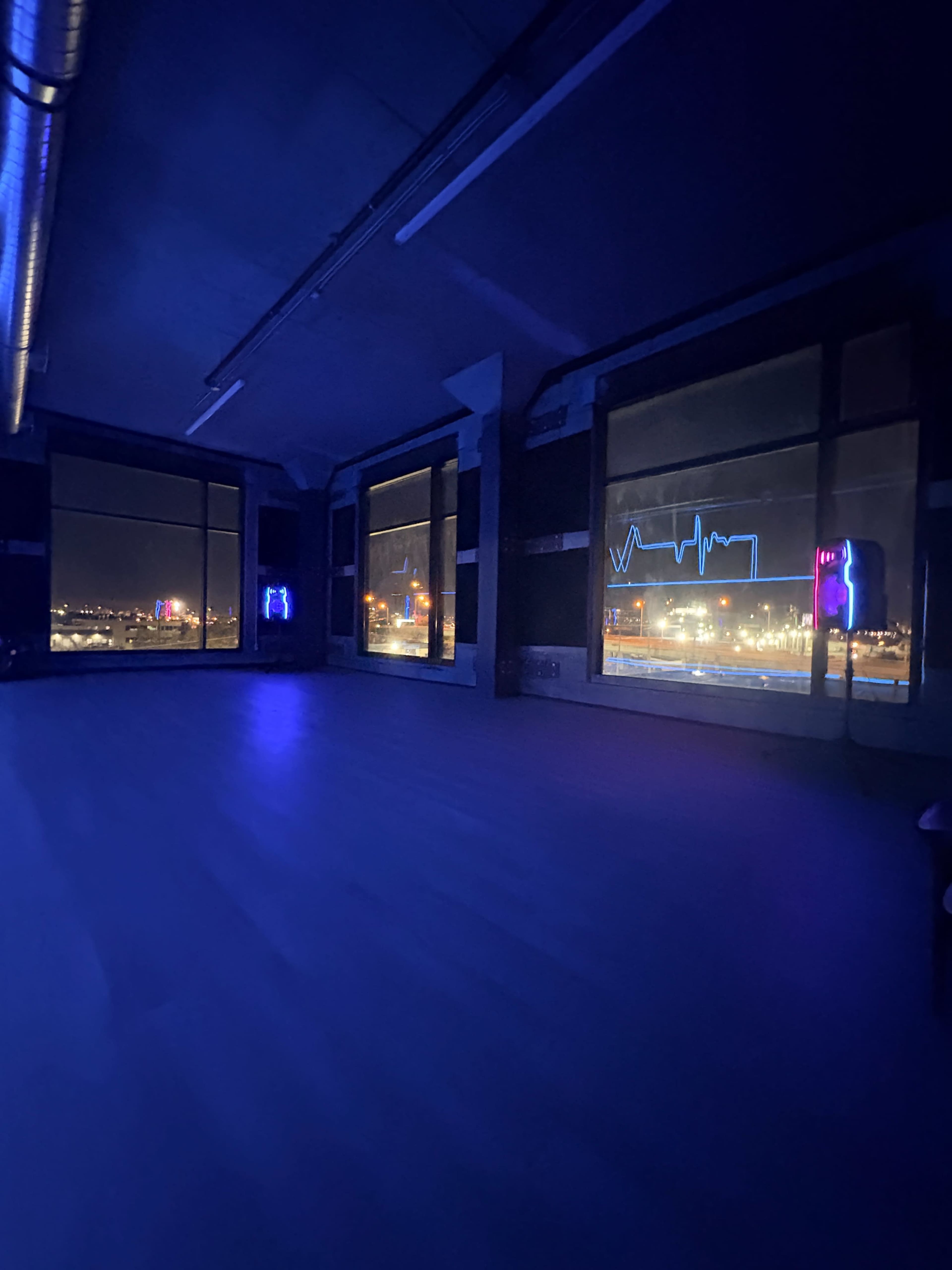 The image shows a dimly lit room with blue lighting and large windows overlooking a city skyline at night, featuring neon signs.