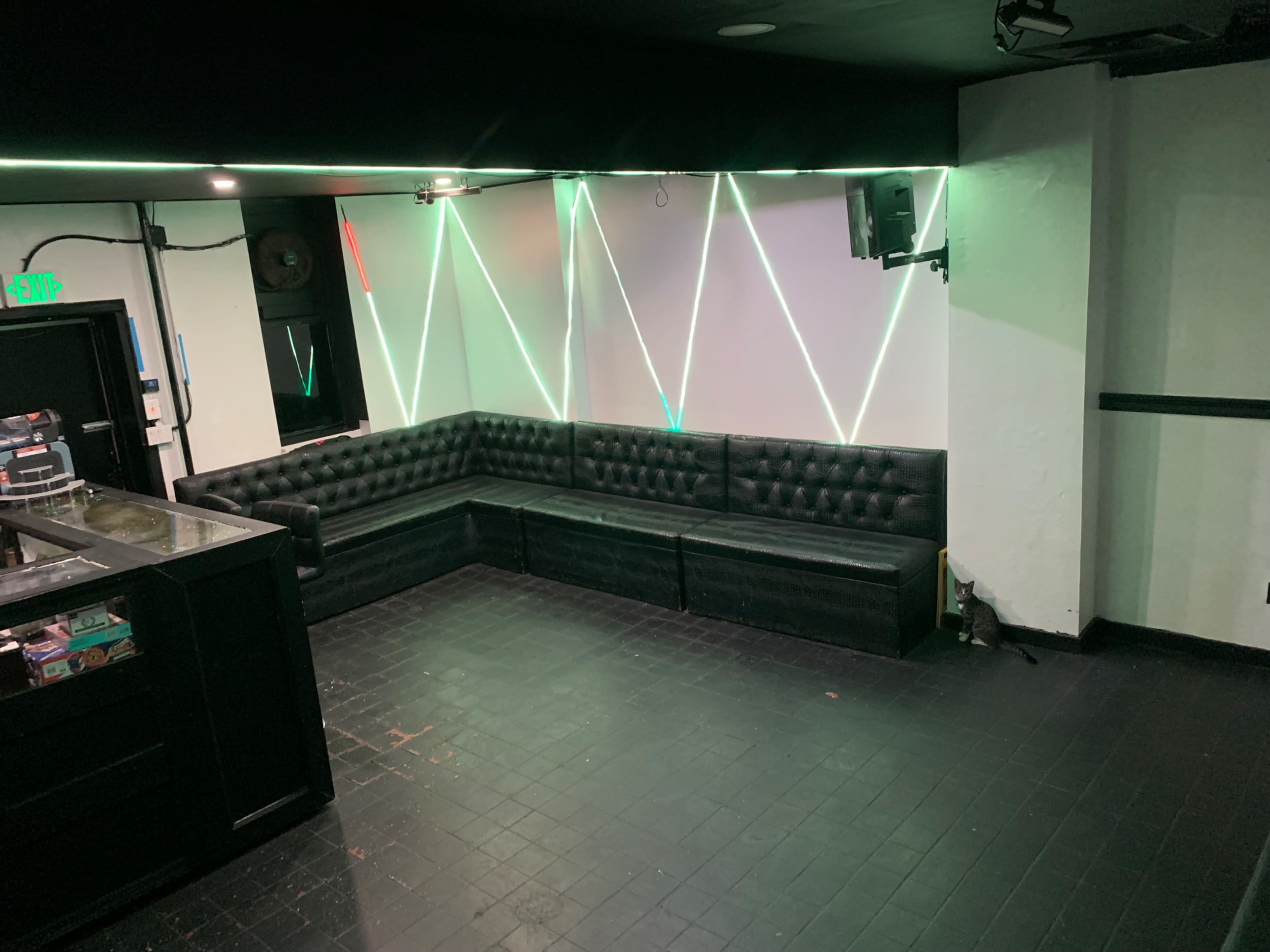 The image shows a dimly lit lounge area with black leather seating arranged along the wall and green and white LED lights creating geometric patterns on the opposite wall.