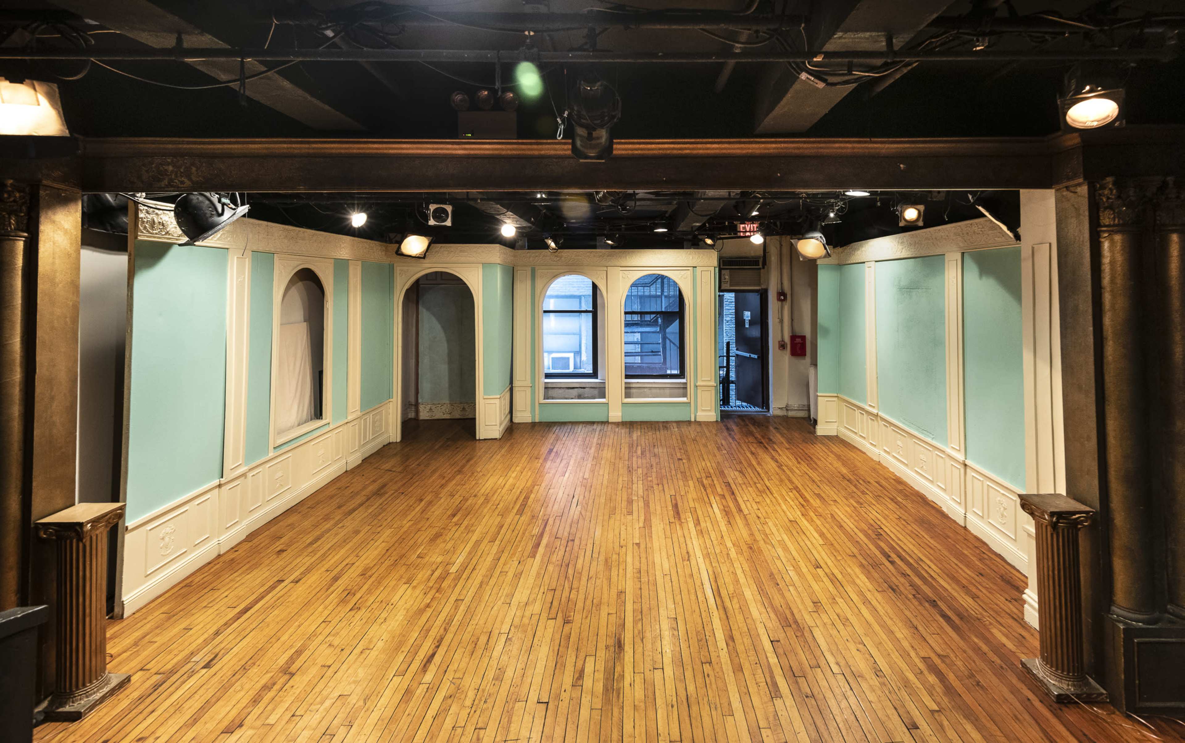 The image shows an empty theater stage with wooden floors, turquoise walls, and arched windows.