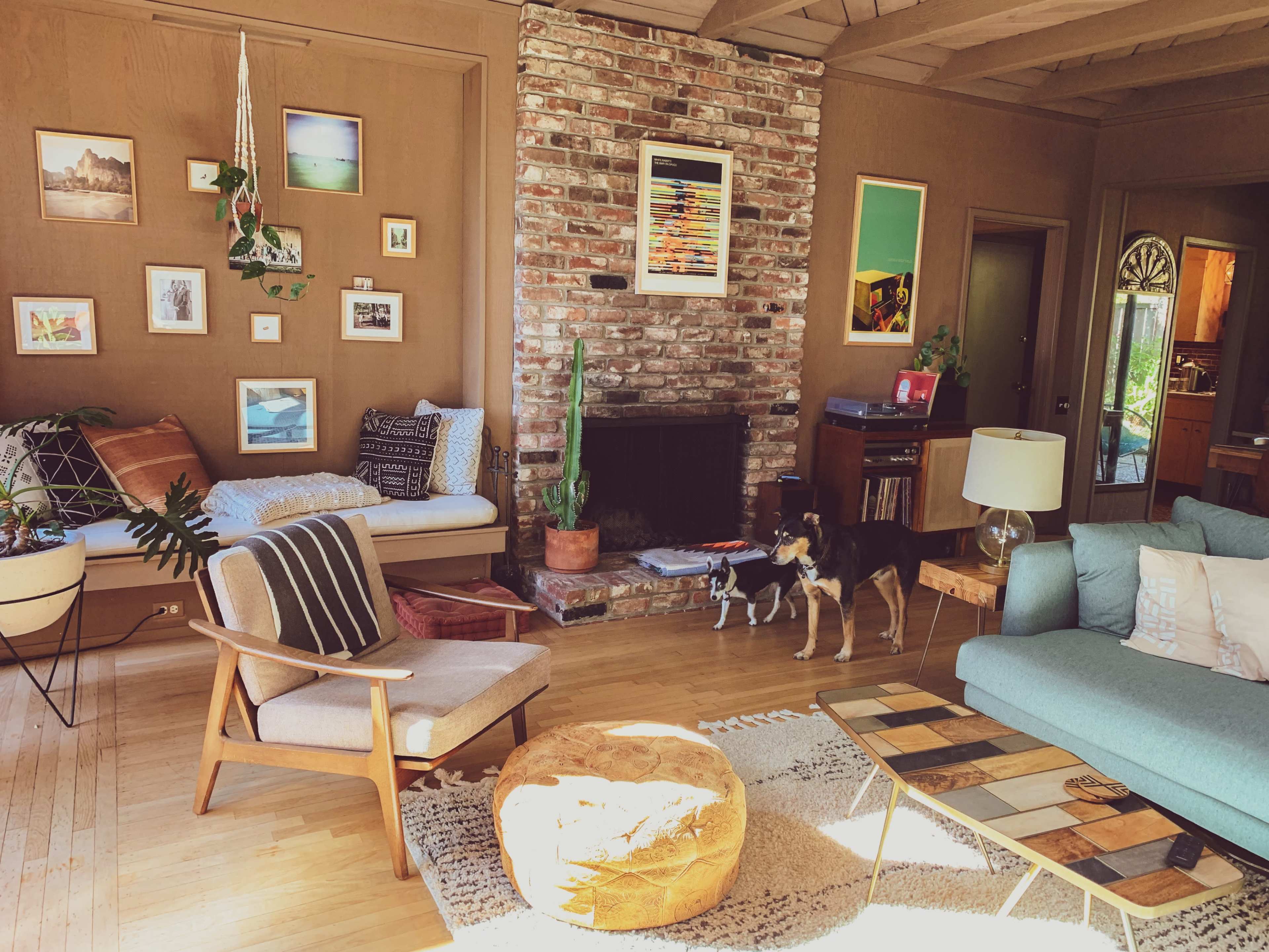 A cozy living room features a brick fireplace, colorful wall art, a green couch, and two dogs playing on the hardwood floor.