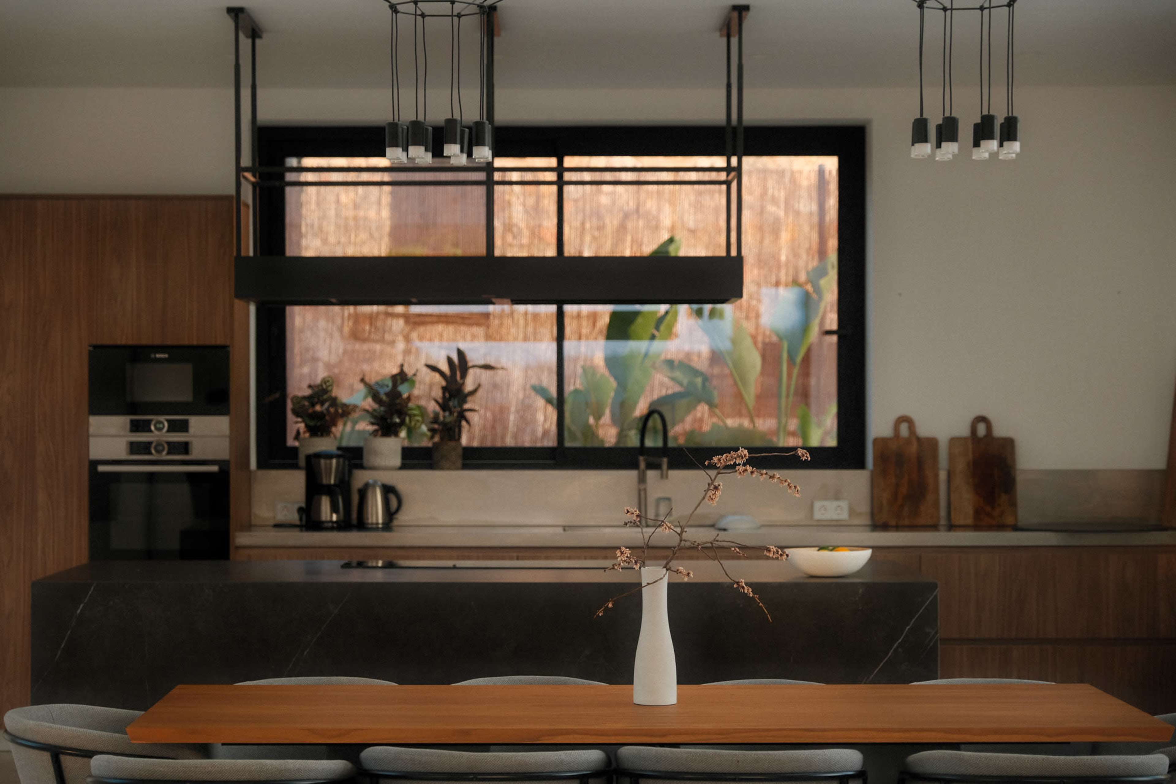 A modern kitchen features a wooden dining table with a vase of branches and a large window showcasing plants outside, complemented by sleek cabinetry and pendant lighting.