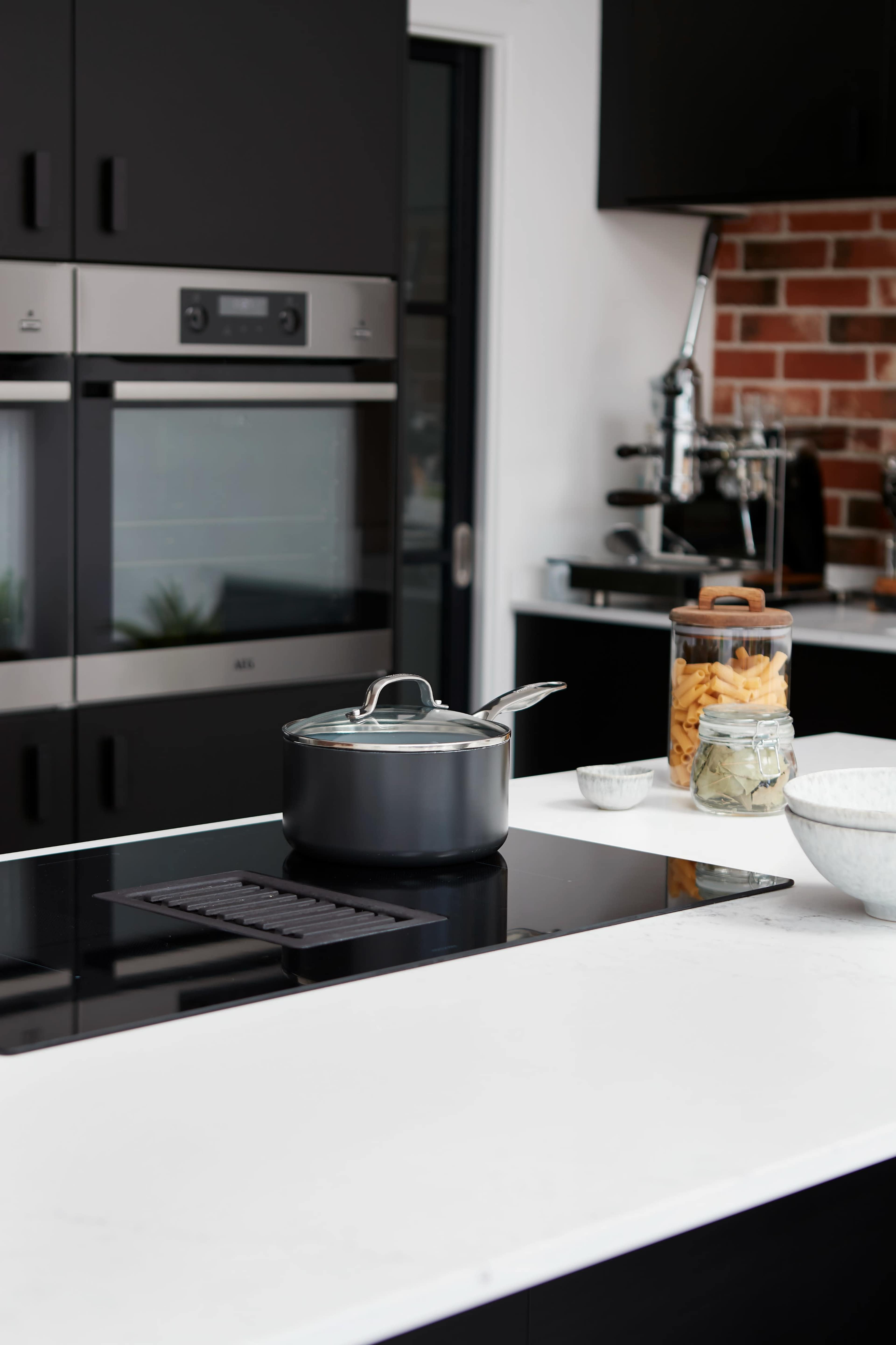 A modern kitchen features a sleek stovetop with a pot, a glass jar of pasta, and an assortment of utensils on a white countertop.