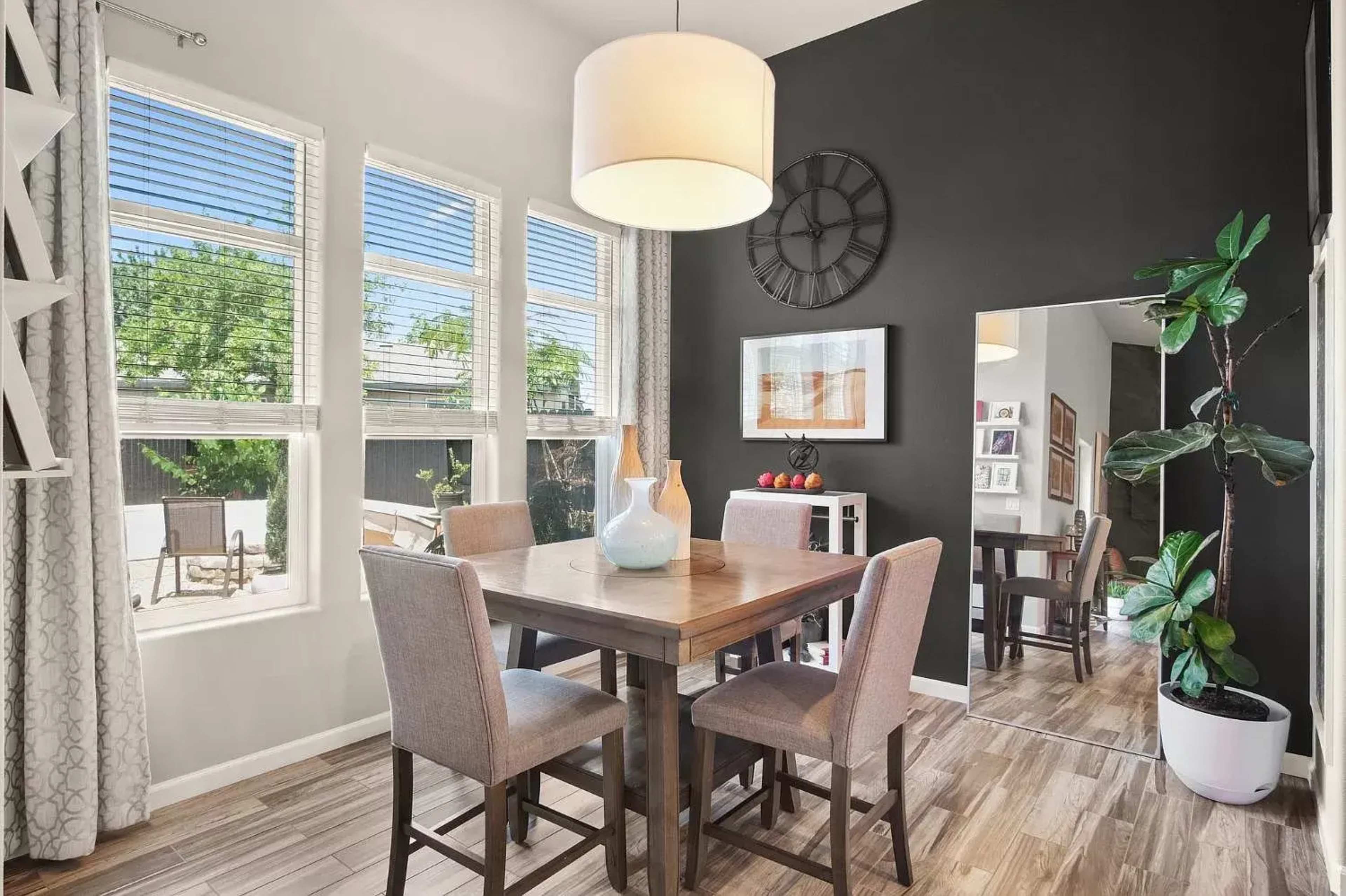 A dining area features a wooden table surrounded by four upholstered chairs, with large windows allowing natural light, and a dark accent wall displaying a clock and mirror.