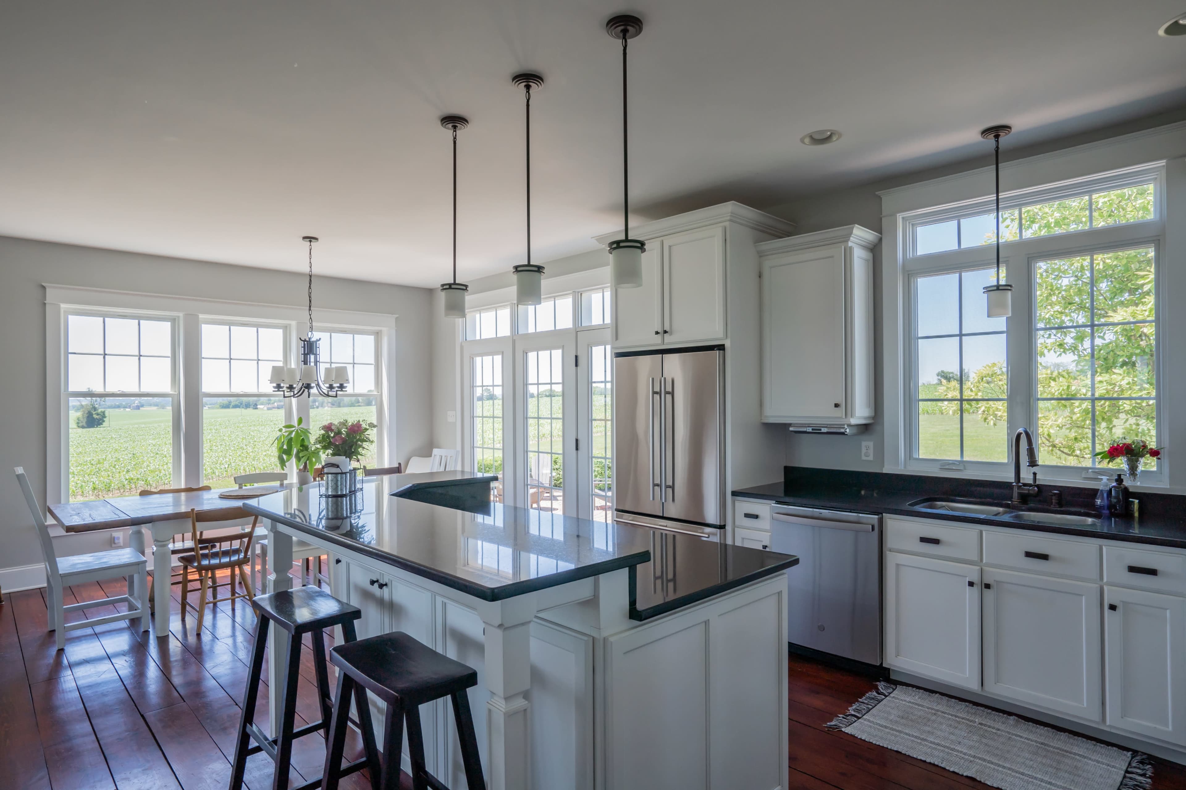 A modern kitchen features white cabinets, a large island with dark countertops, and dining space with a view of the landscape through expansive windows.
