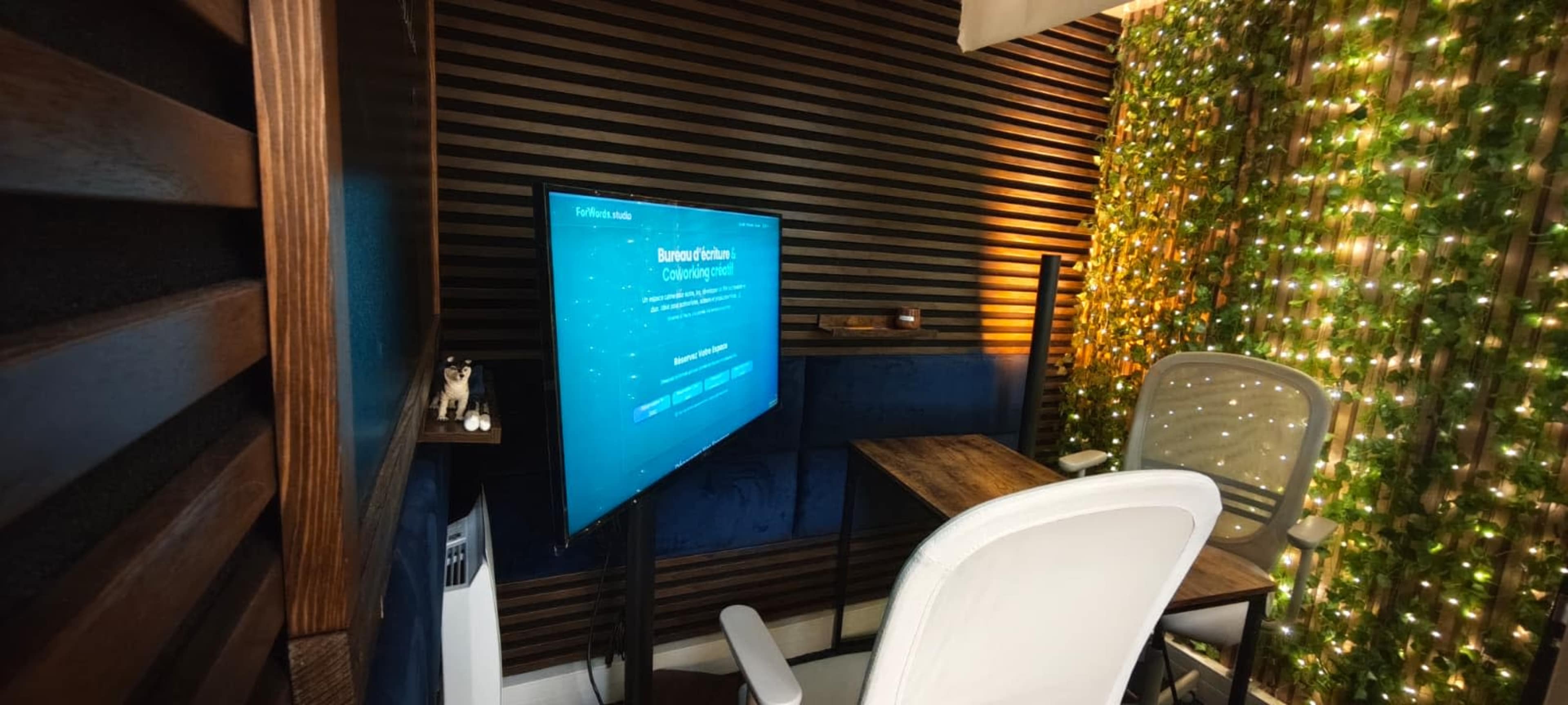 A modern workspace features a wooden desk, a white office chair, and a television mounted on the wall, surrounded by a green backdrop of decorative lights.