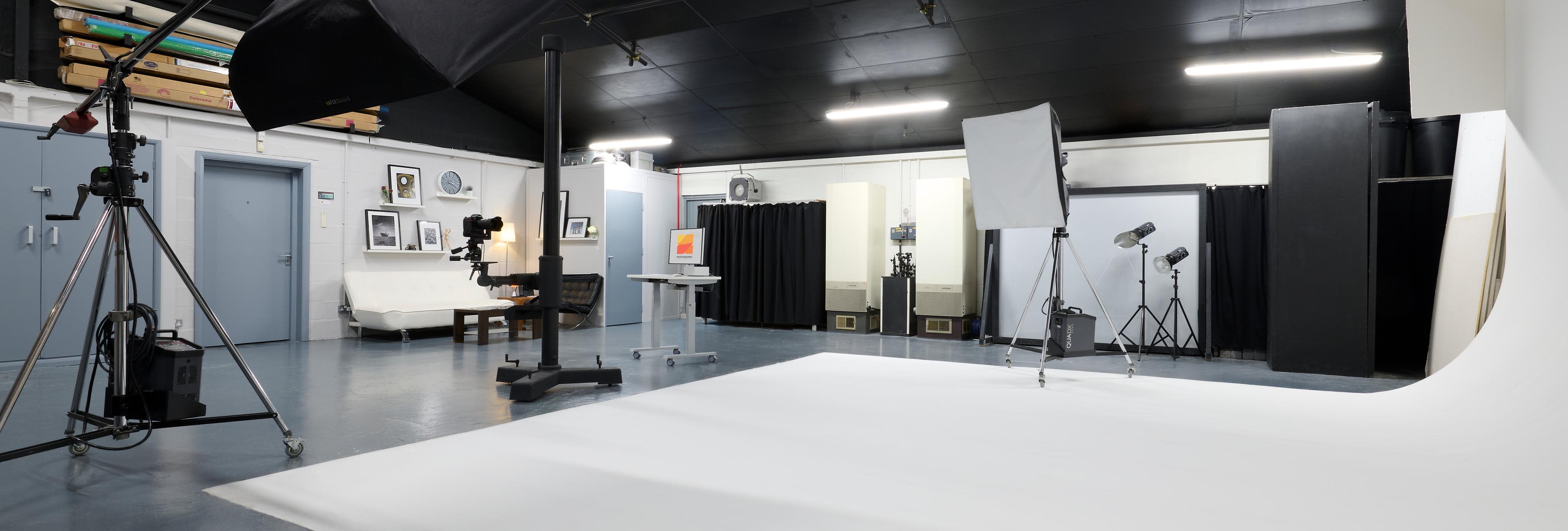 The image depicts a spacious photography studio featuring various lighting equipment, a seamless backdrop, and a seating area.