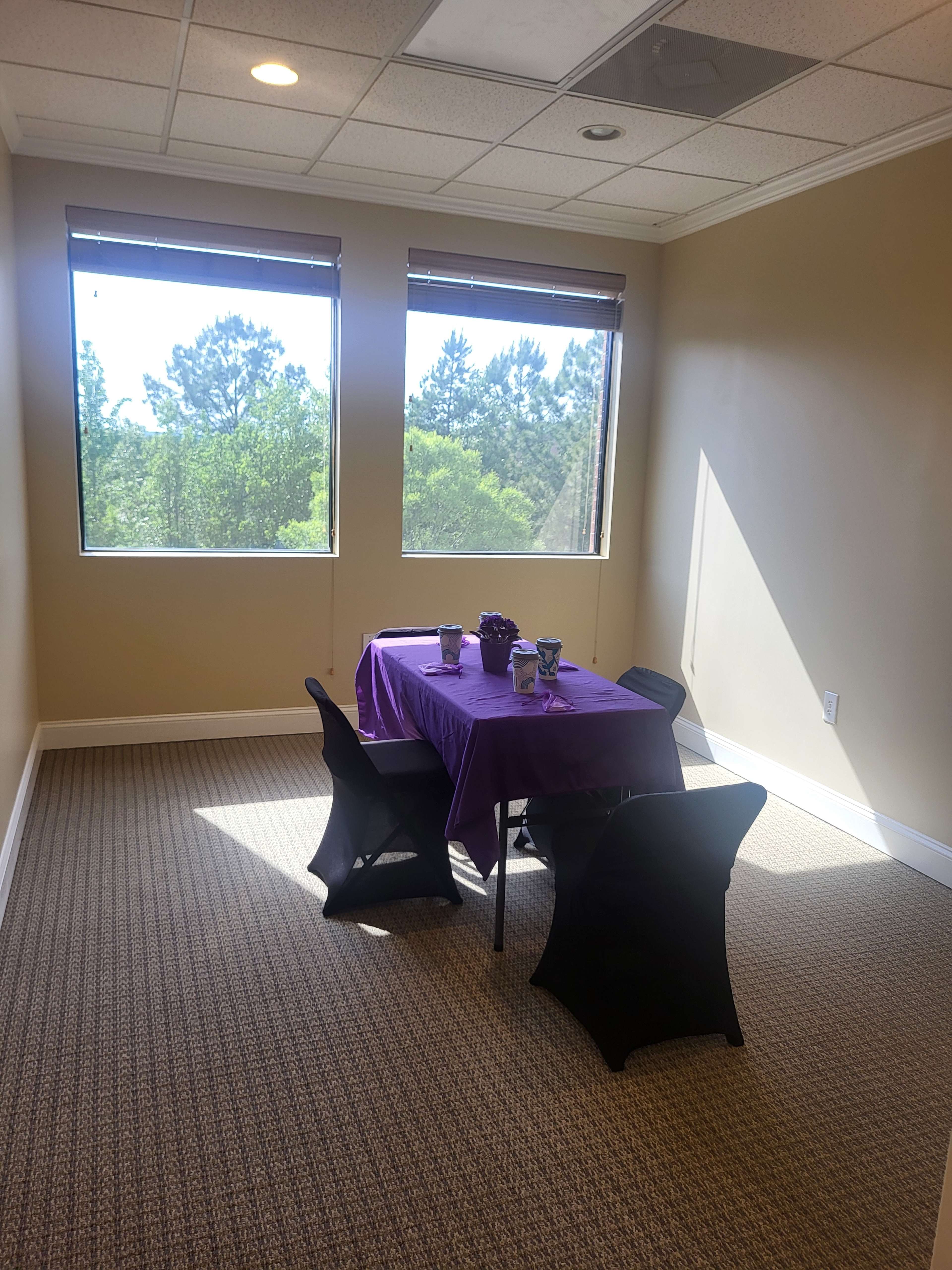 A small room features a table covered with a purple cloth and four black chairs, illuminated by natural light from two windows.