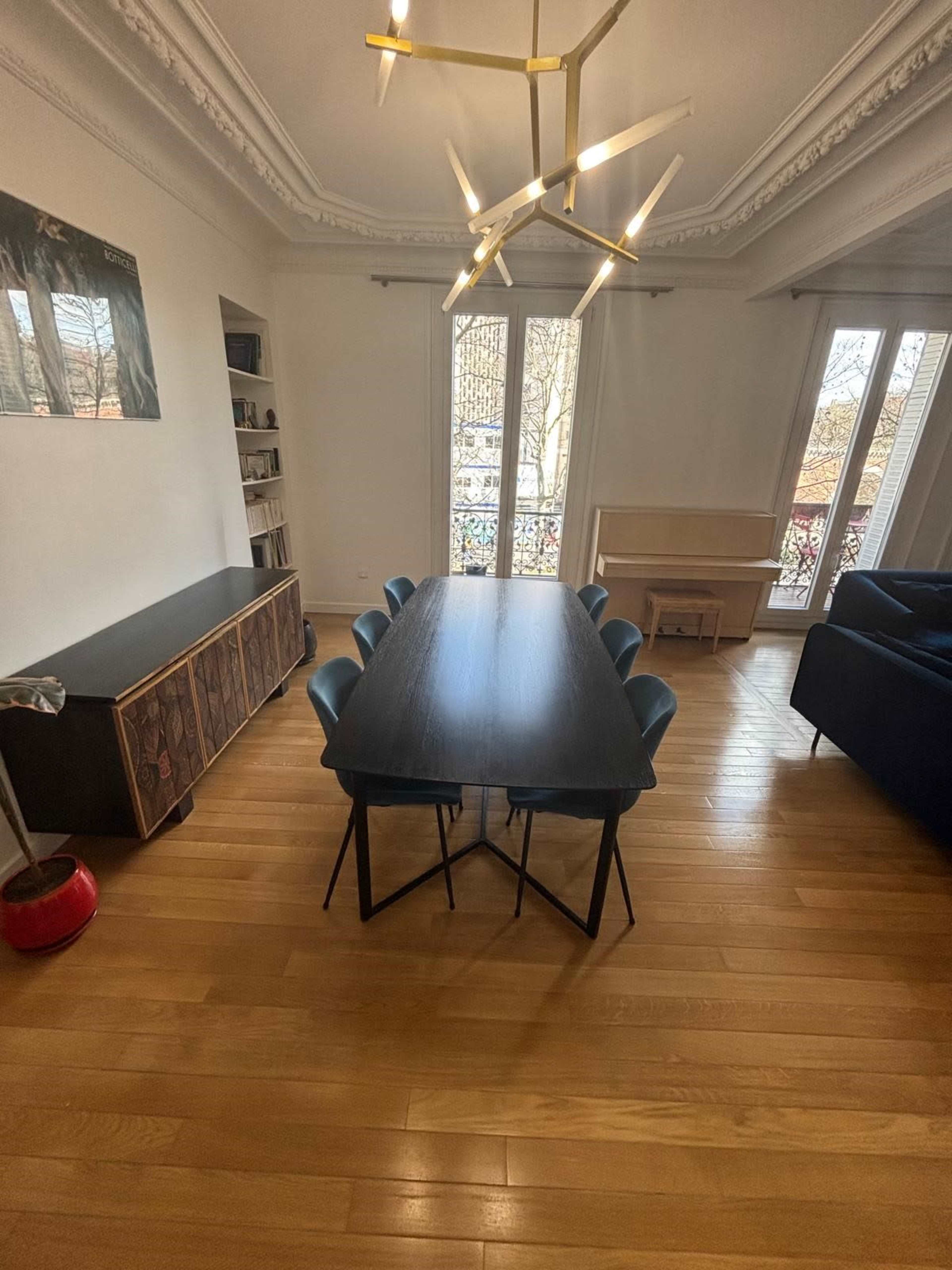 The image shows a modern dining area with a long black table surrounded by six blue chairs, hardwood flooring, and large windows displaying a view of a balcony.