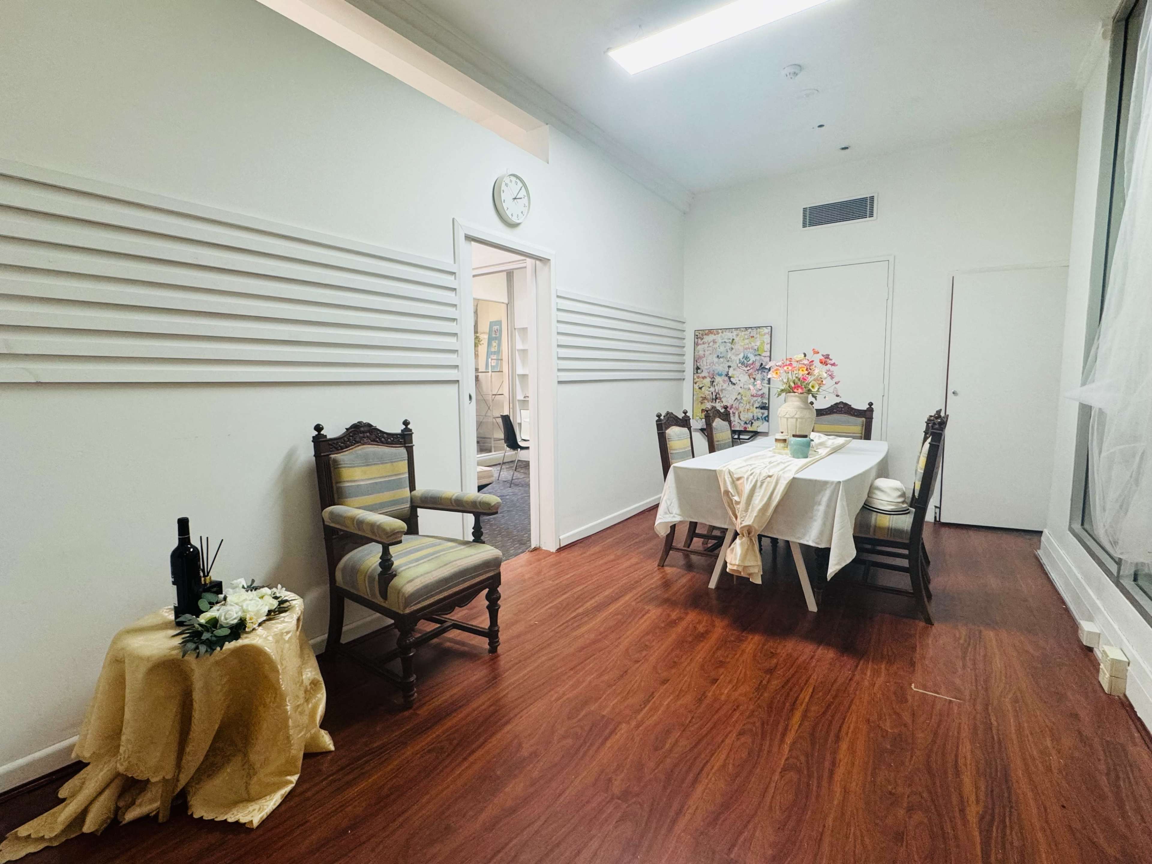 The image shows a sparsely decorated room with a dining table set for a meal, surrounded by chairs, and a small table with a decorative arrangement in one corner.