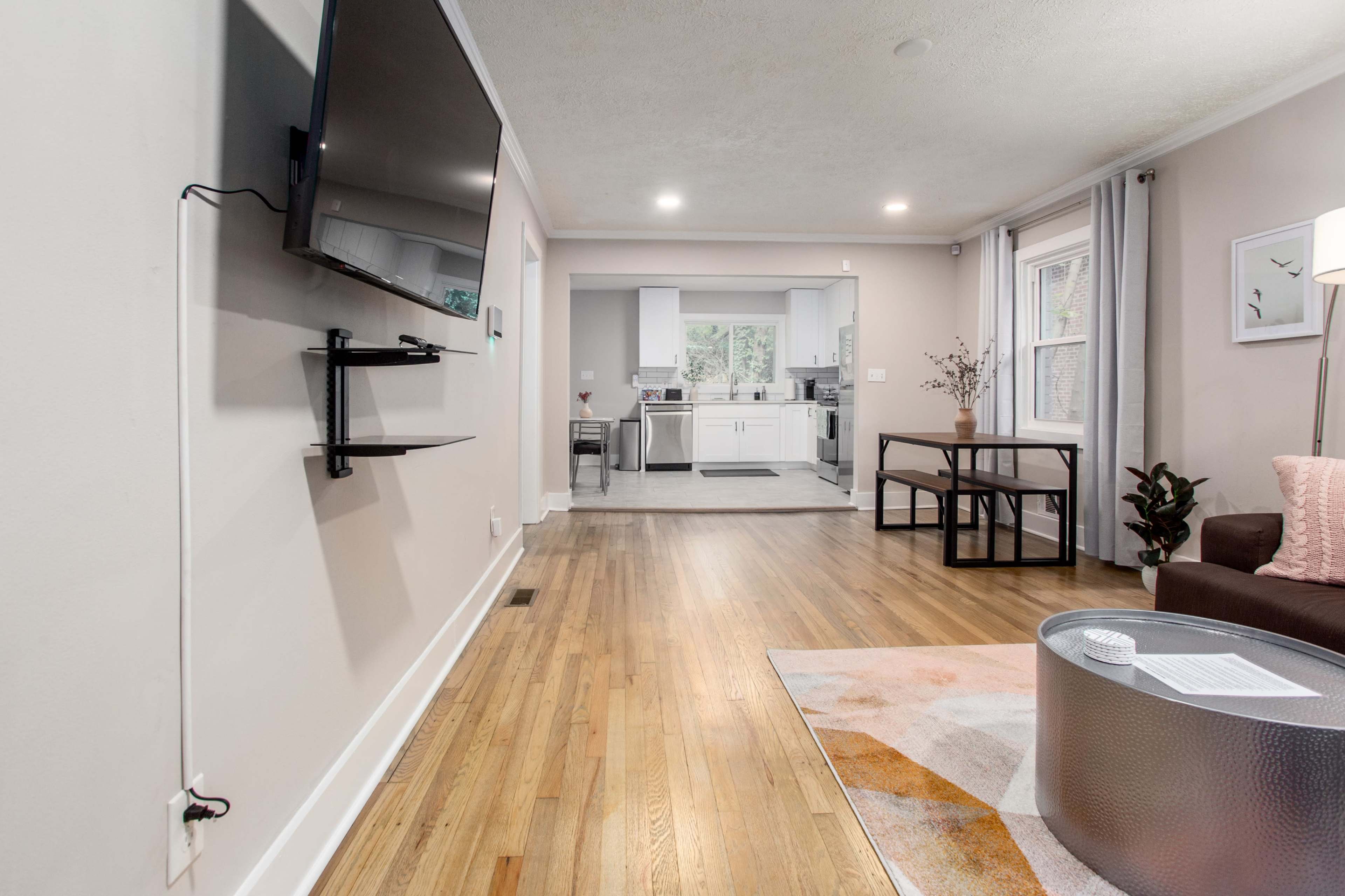 A living room with a wall-mounted television and a view into a kitchen area, featuring hardwood floors and a small dining table.