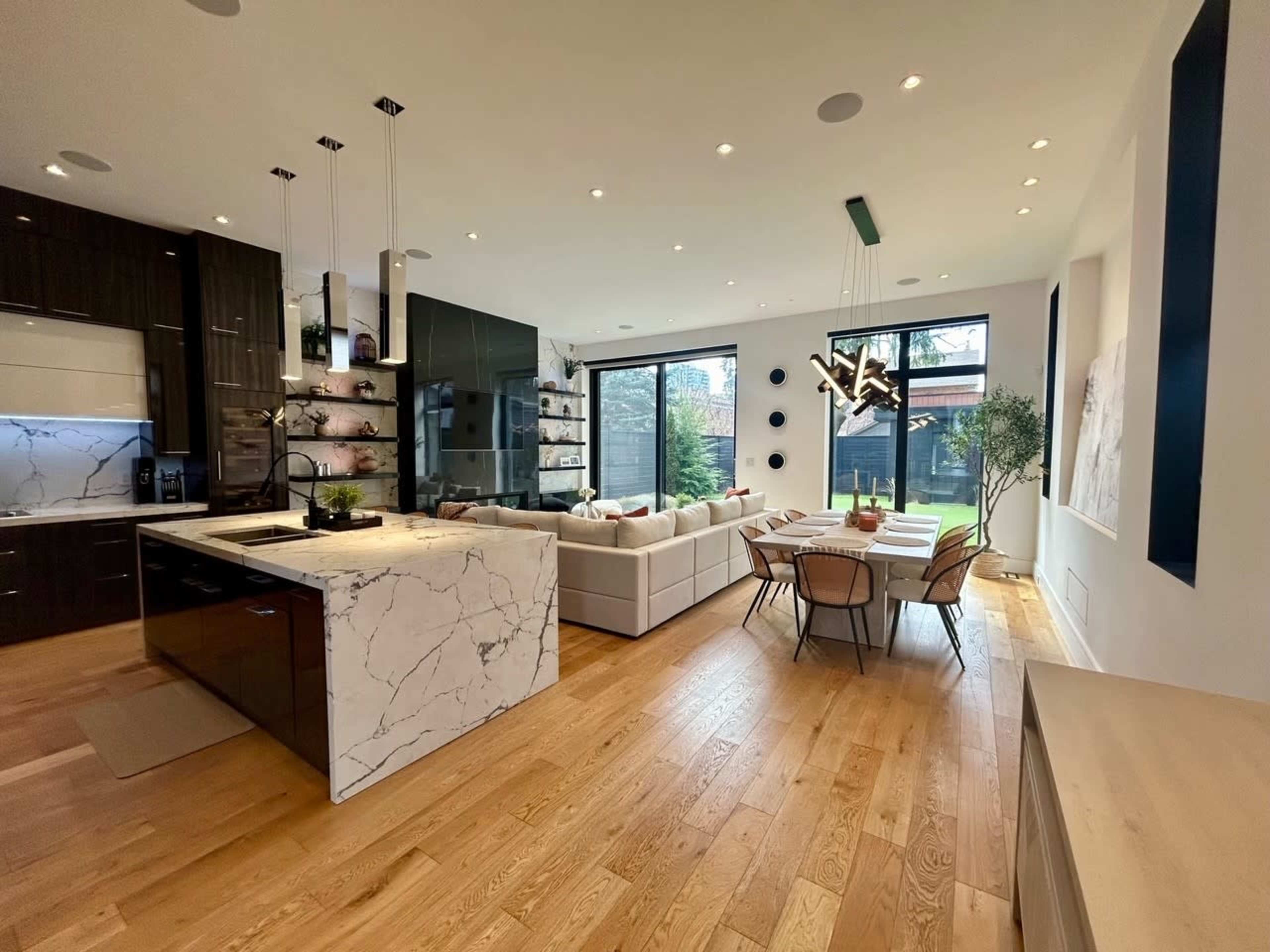 A modern kitchen and dining area feature sleek black cabinets, a marble-topped island, a spacious living area with a couch, and large windows overlooking a garden.
