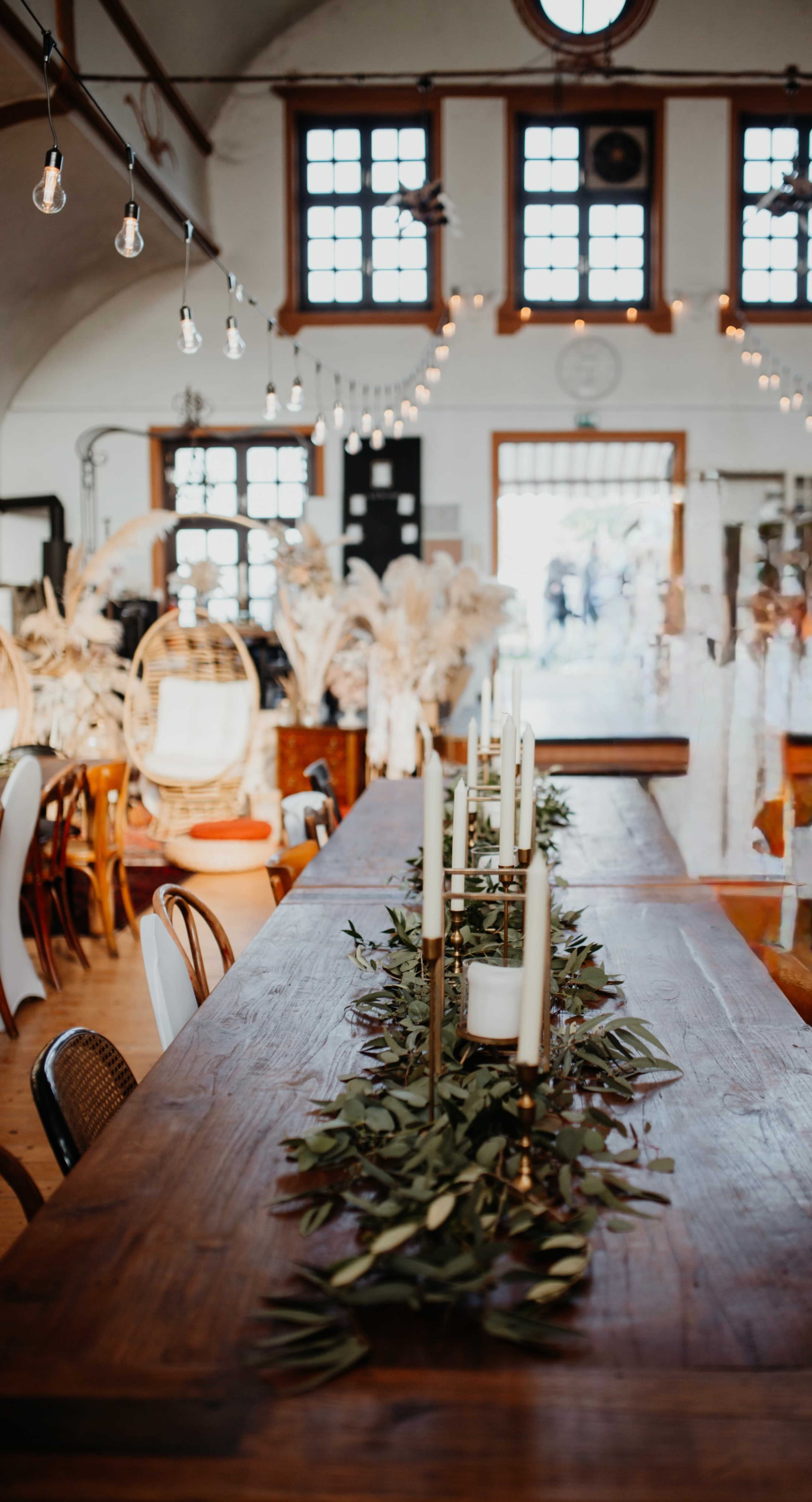 The image shows a dining space with a long wooden table decorated with greenery and candles, surrounded by various chairs and light fixtures.