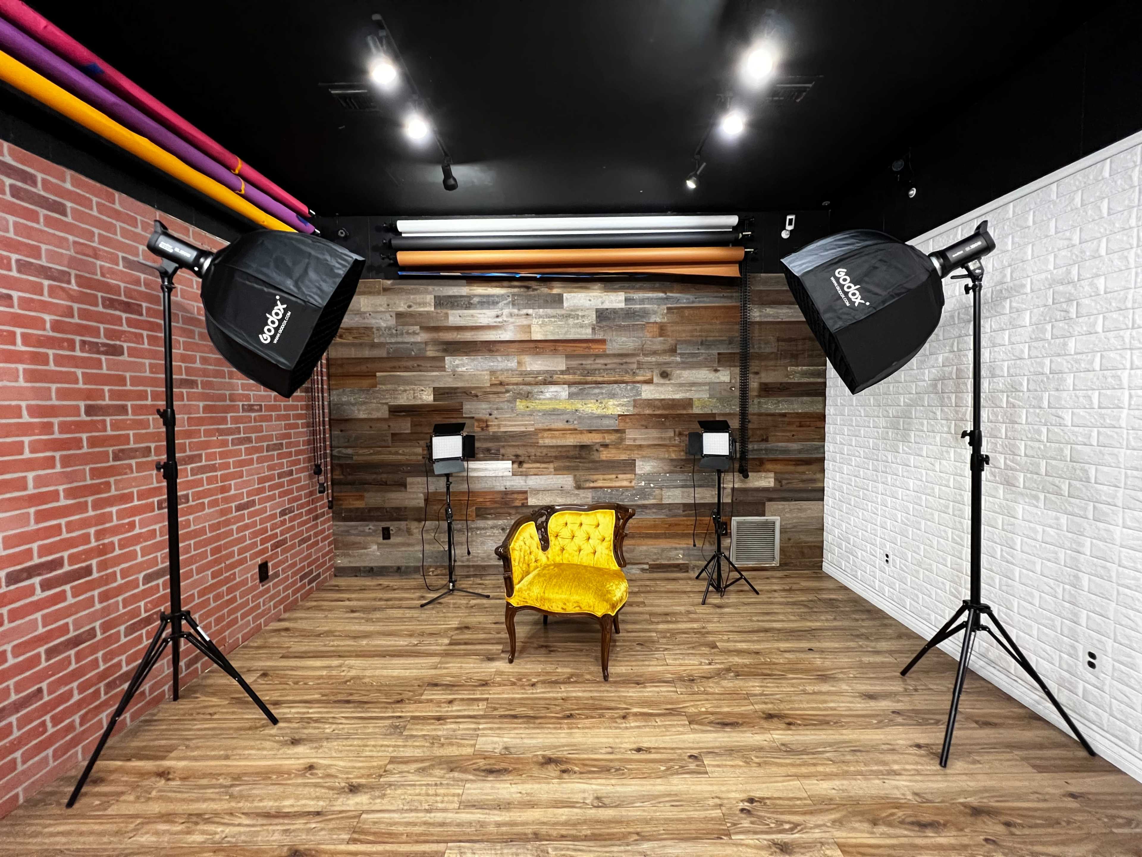 A photography studio featuring a yellow upholstered chair positioned in the center, flanked by two softbox lighting setups against a backdrop of textured wooden panels and white brick walls.