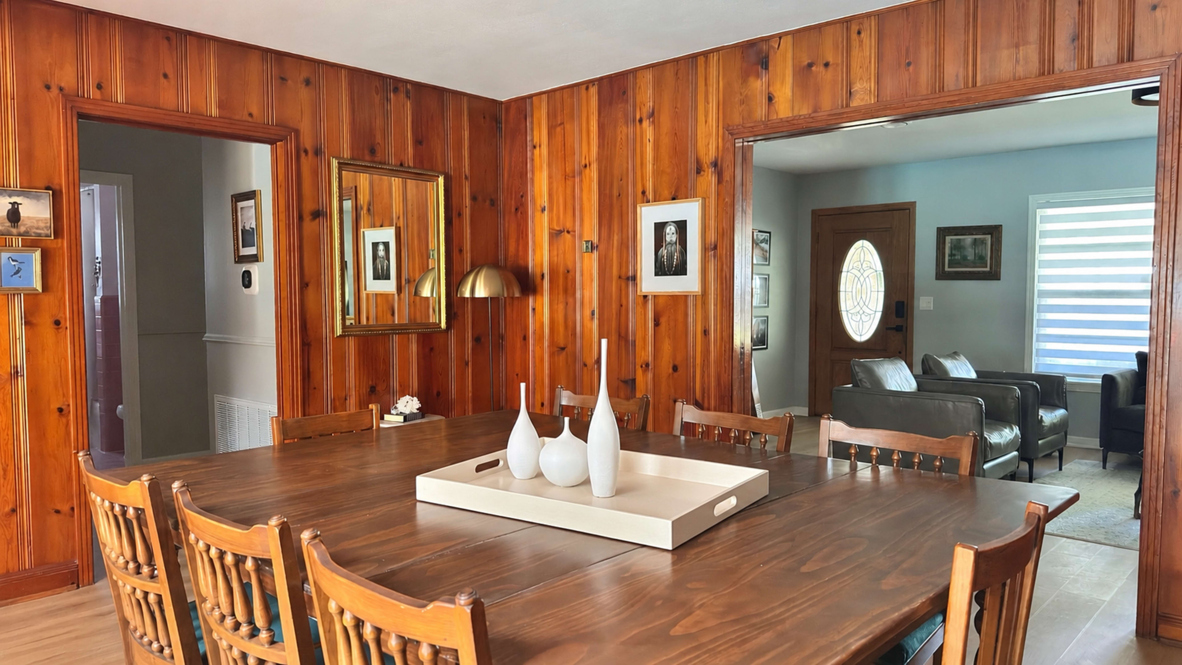 The image shows a wooden dining room with a table set for eight, featuring a central tray with decorative vases, and walls adorned with wooden paneling and framed pictures.
