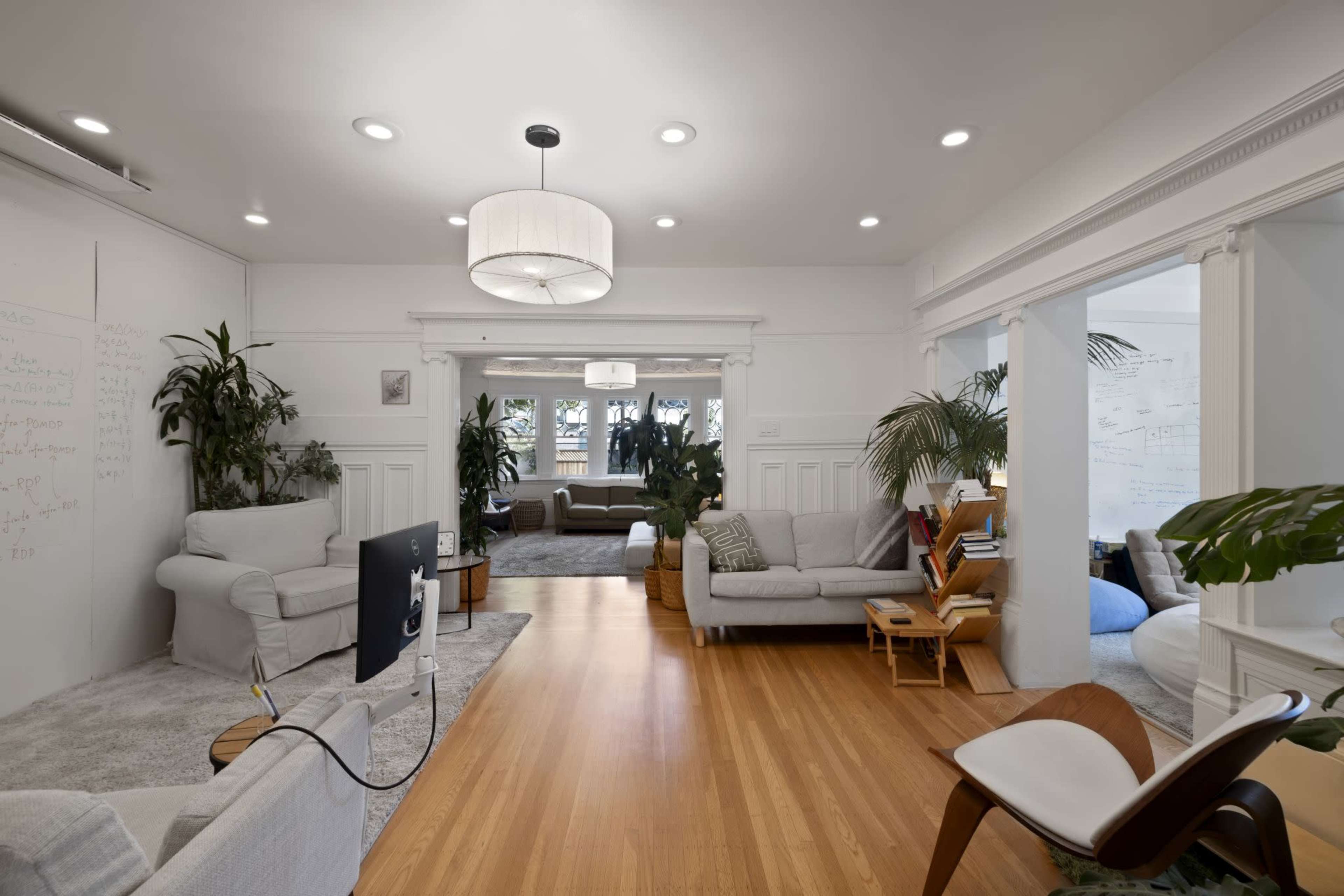 The image shows a bright living room featuring white walls, hardwood floors, and a mix of comfortable seating, plants, and a television.